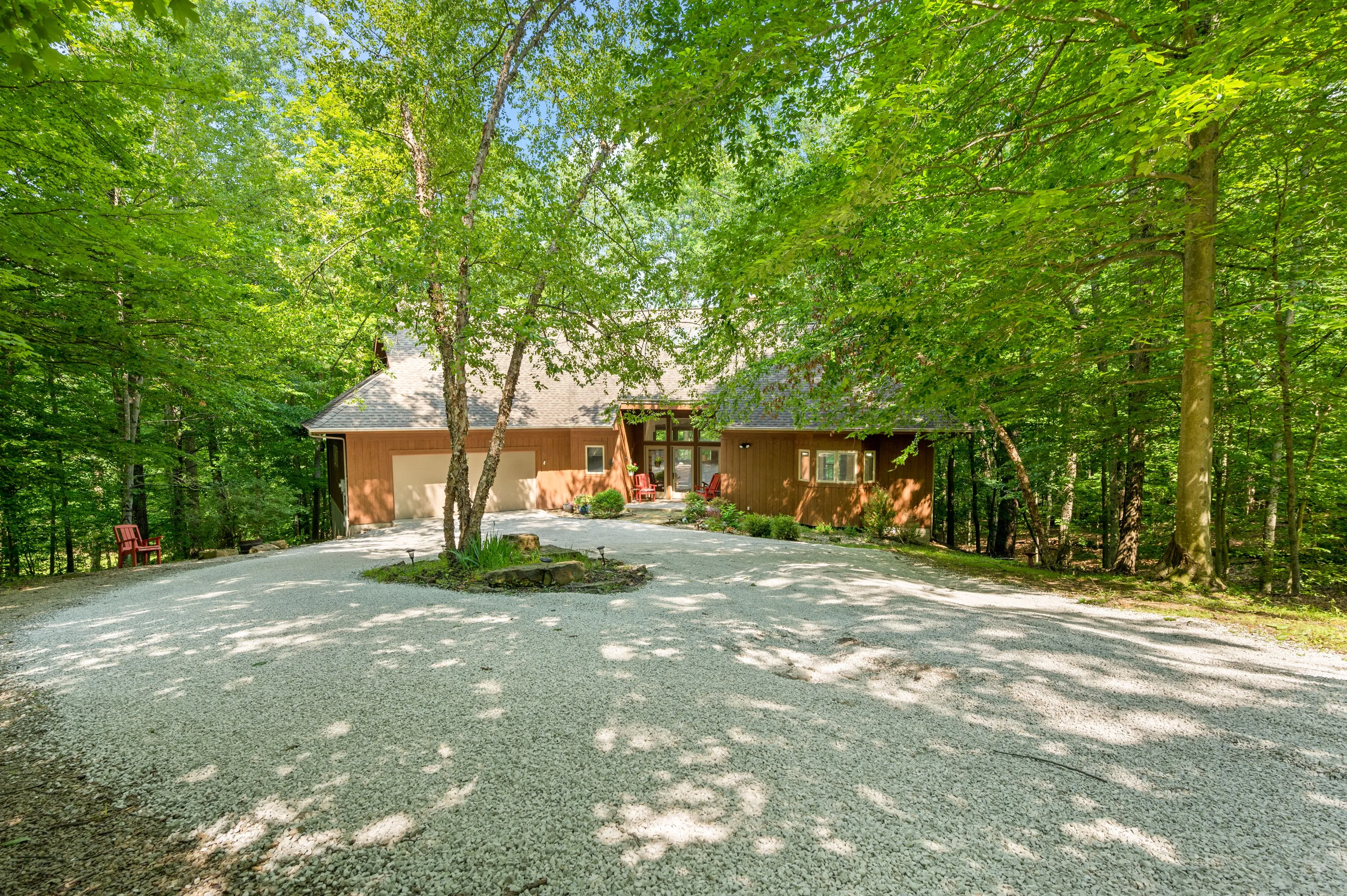 Brick house nestled in lush greenery with a circular driveway and scattered sunlight through the trees.