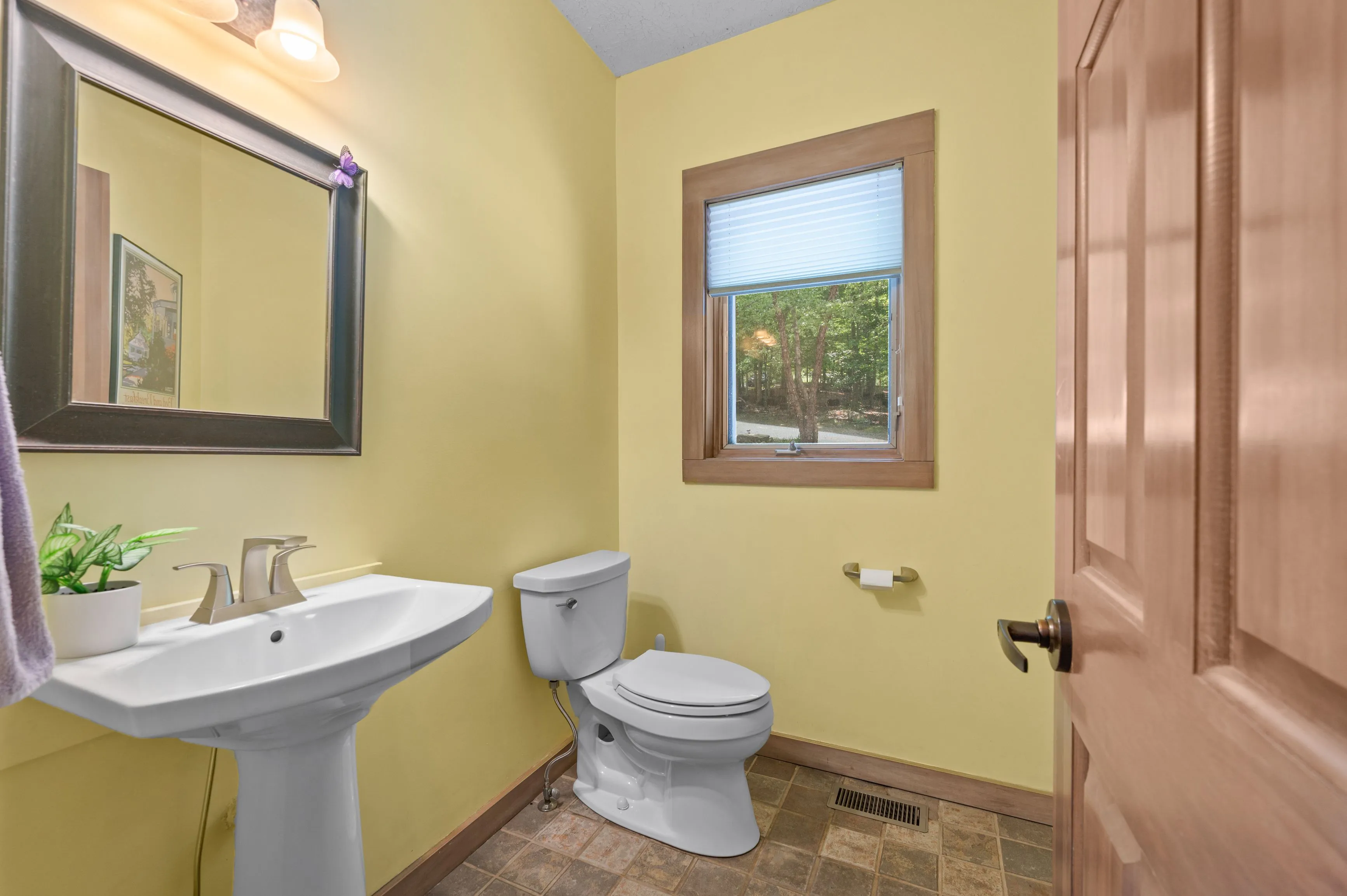 Interior of a small bathroom with yellow walls, white pedestal sink, toilet, and a shower curtain partially visible to the right. A window provides natural light.