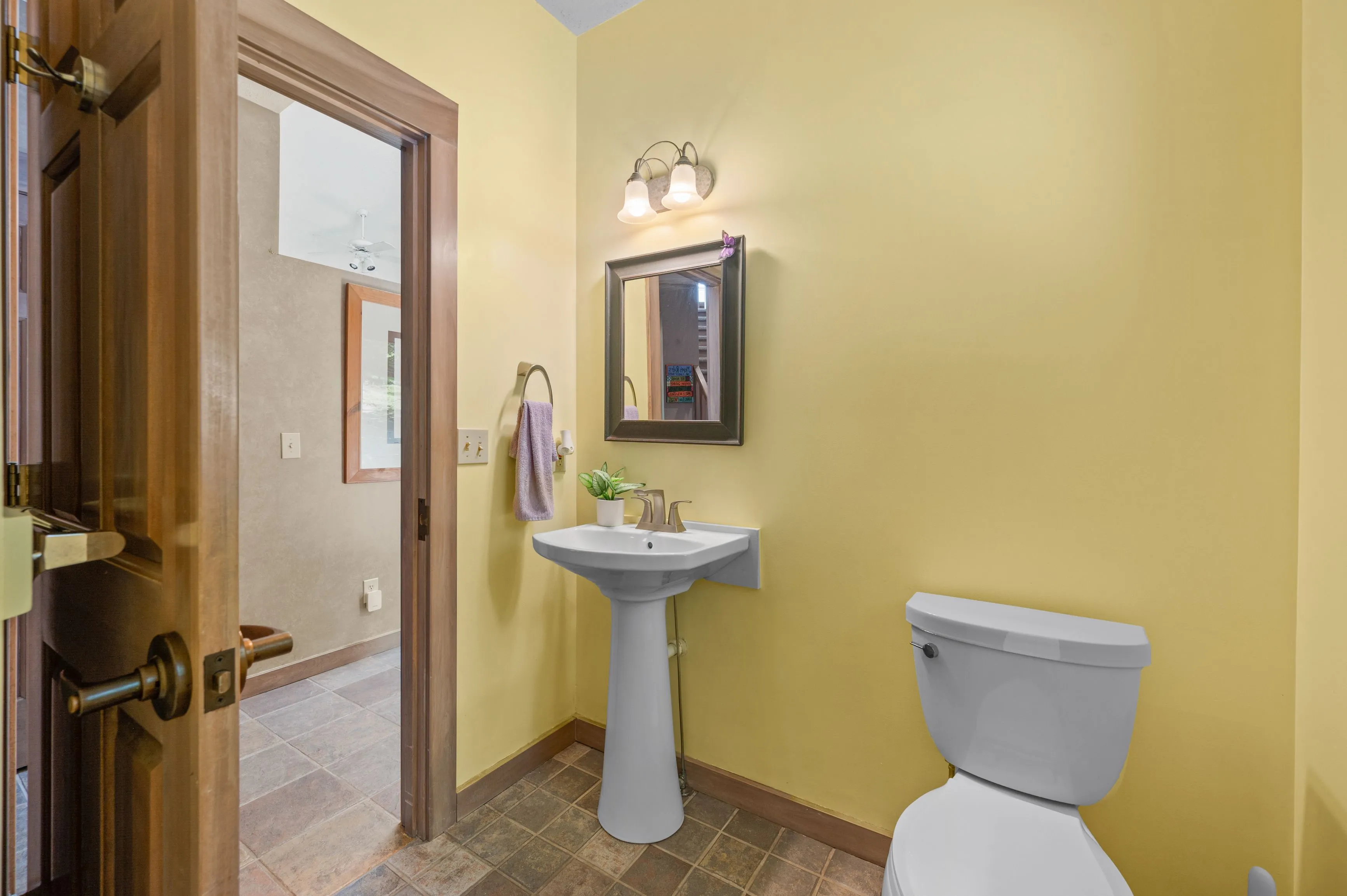 A small bathroom with yellow walls, featuring a pedestal sink, toilet, and a framed mirror with decorative lighting above it.