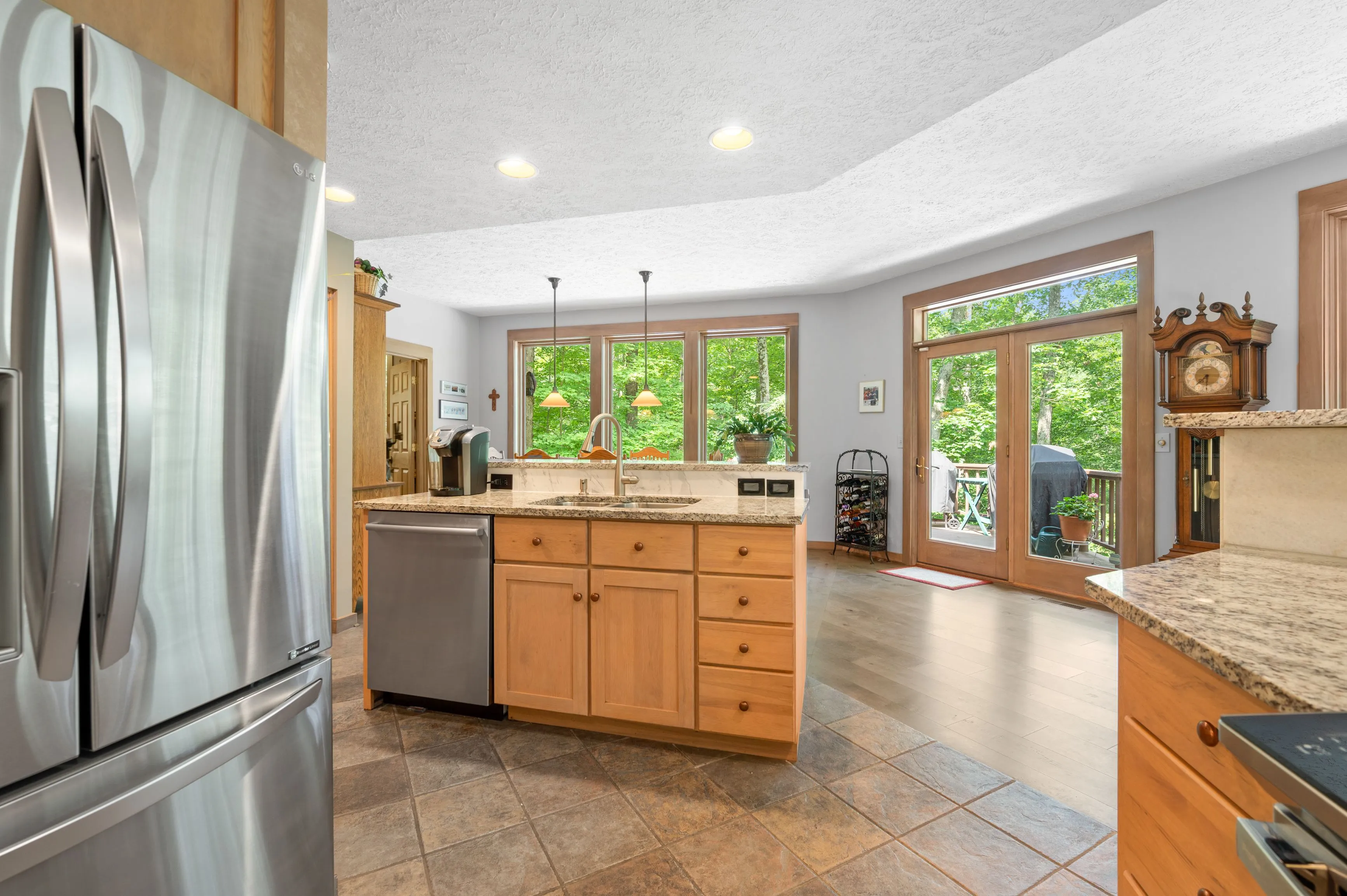 Bright modern kitchen with stainless steel refrigerator, wooden cabinets, and a view of greenery outside the window.