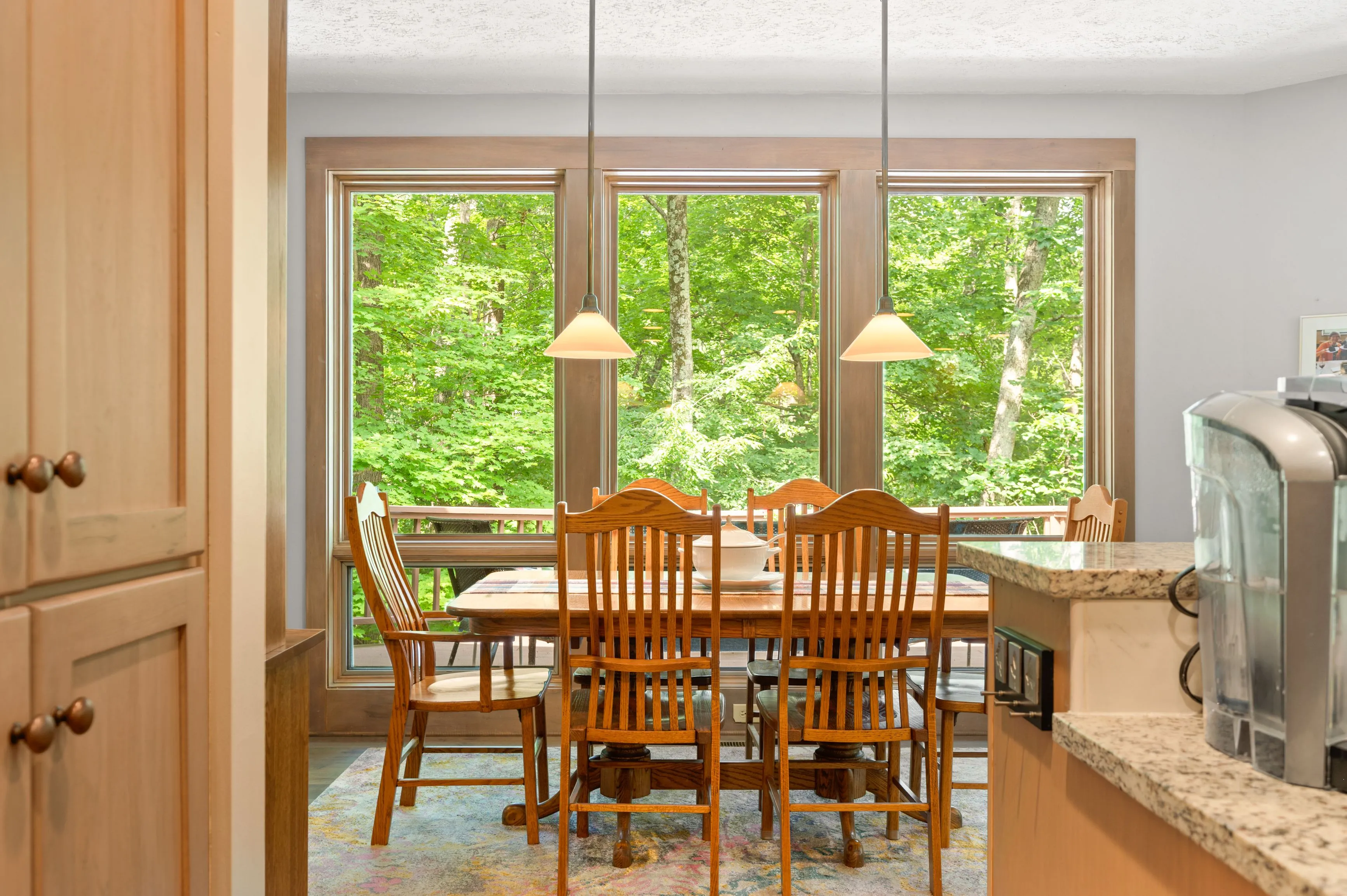 Cozy breakfast nook with wooden table and chairs, pendant lights, surrounded by large windows with a green forest view.