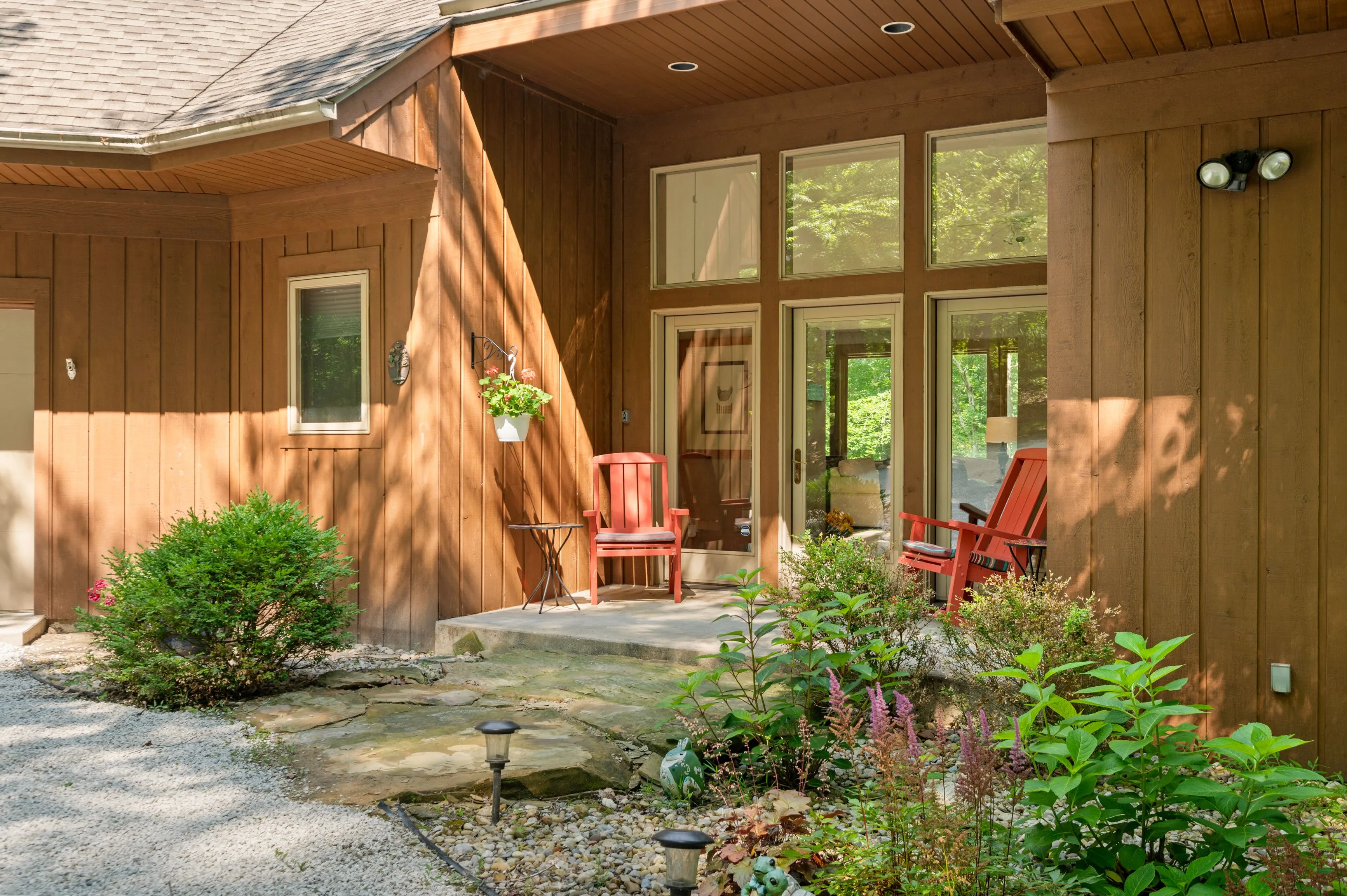 Exterior view of a cozy woodland home with a stone pathway, large windows, and red chairs on the porch surrounded by greenery.