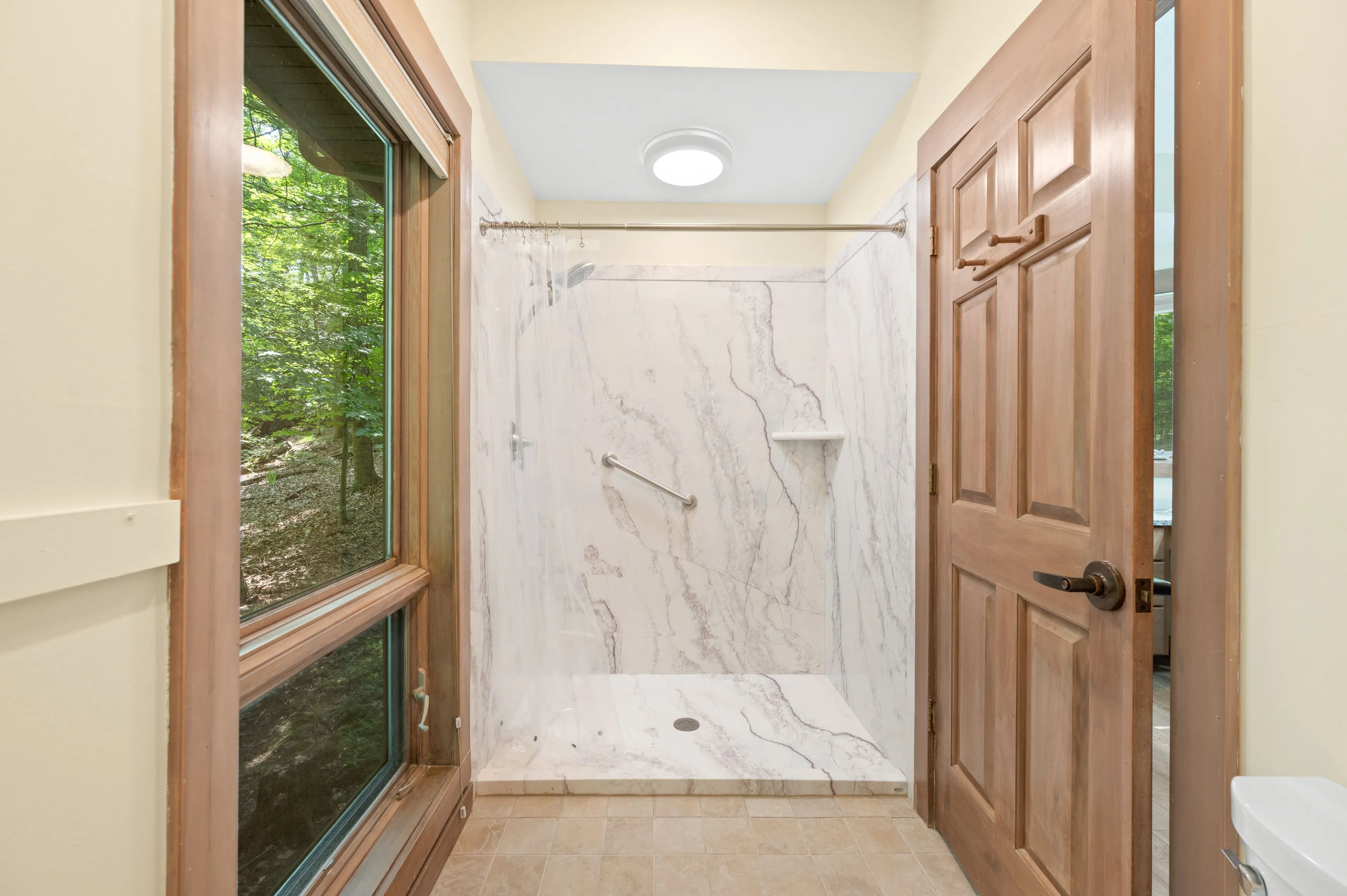 Interior of a modern bathroom with a large walk-in shower, wooden doors, and a window overlooking greenery.