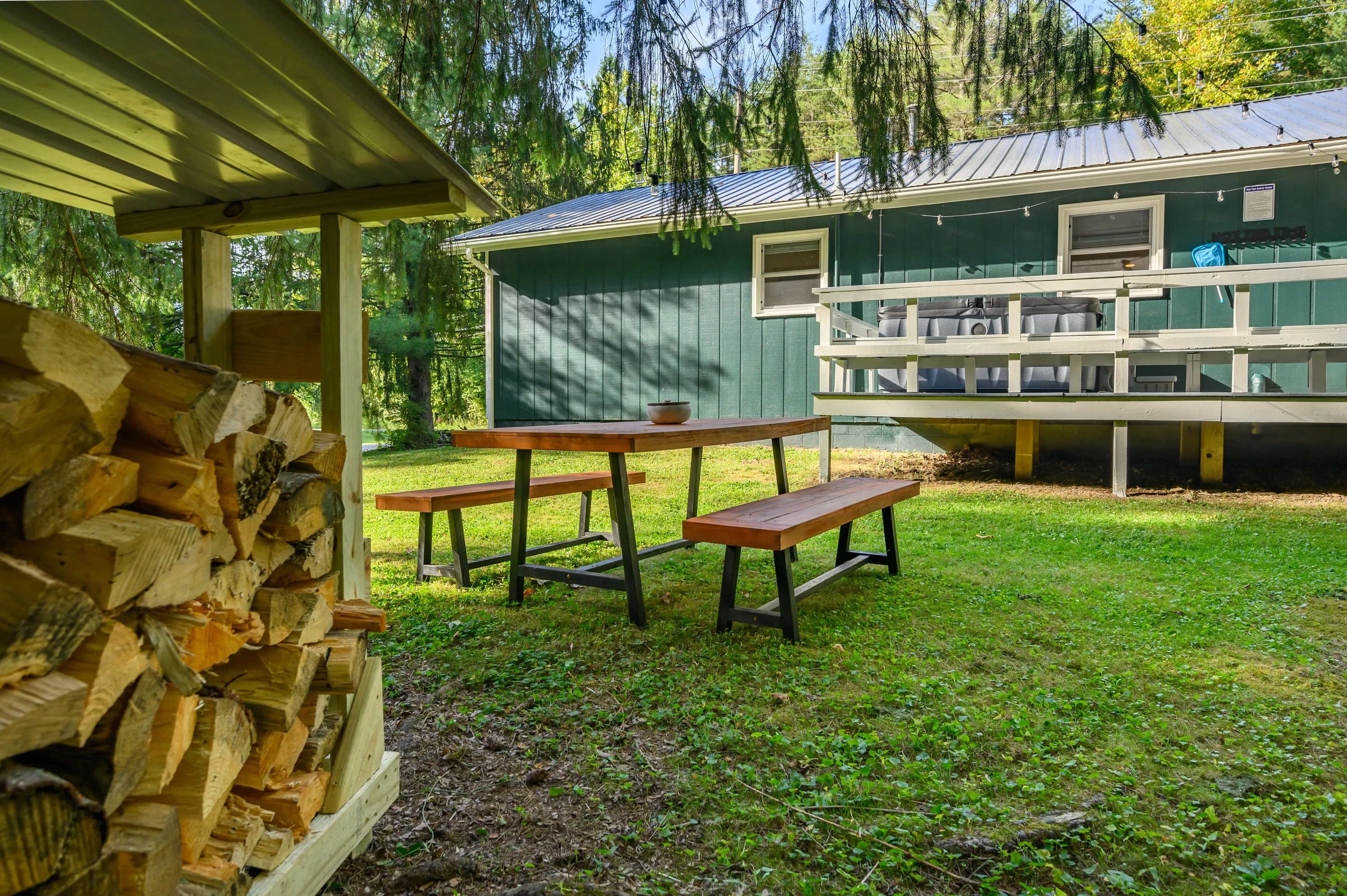 Wooden picnic table and pile of chopped firewood next to a green cabin with a porch in a forested area.