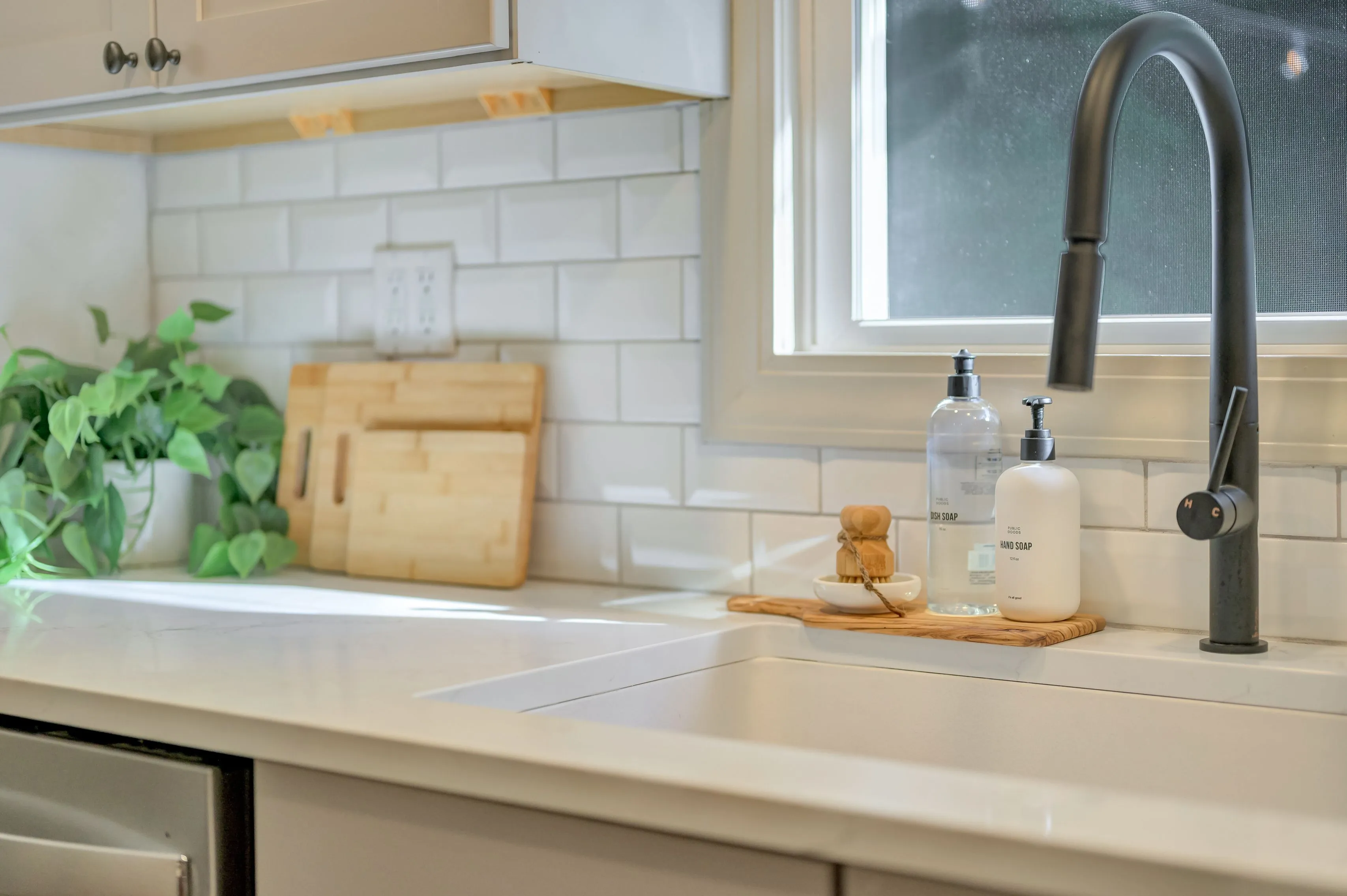 Modern kitchen interior with white countertops, subway tile backsplash, and a black faucet, also showing dish soap on a tray, cutting boards, and a potted plant.