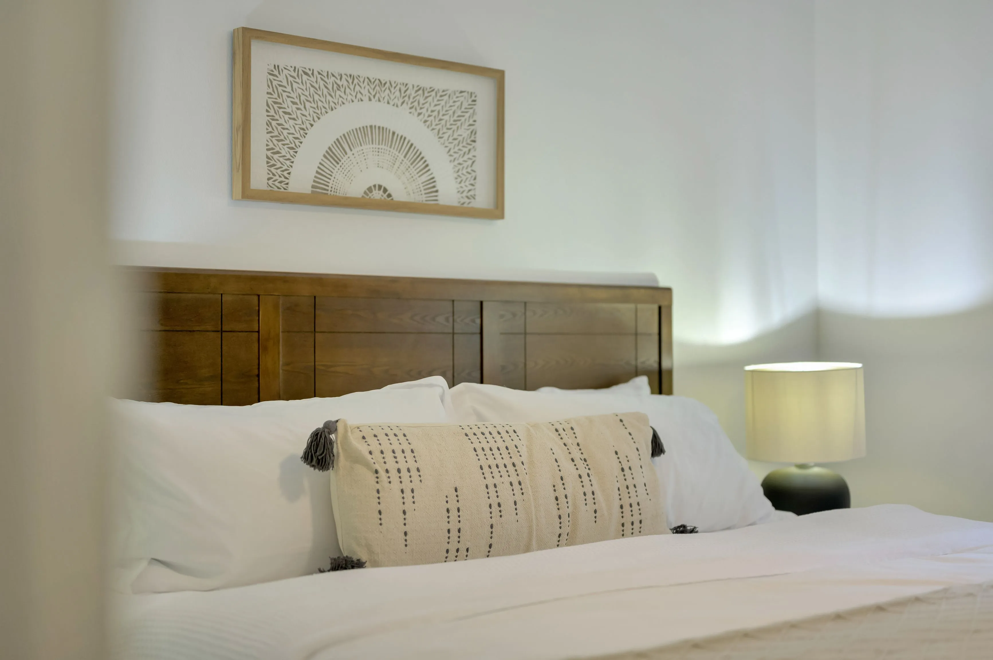 Elegantly made bed with white linens, decorative pillows, wooden headboard, artwork above the bed and a lit table lamp.
