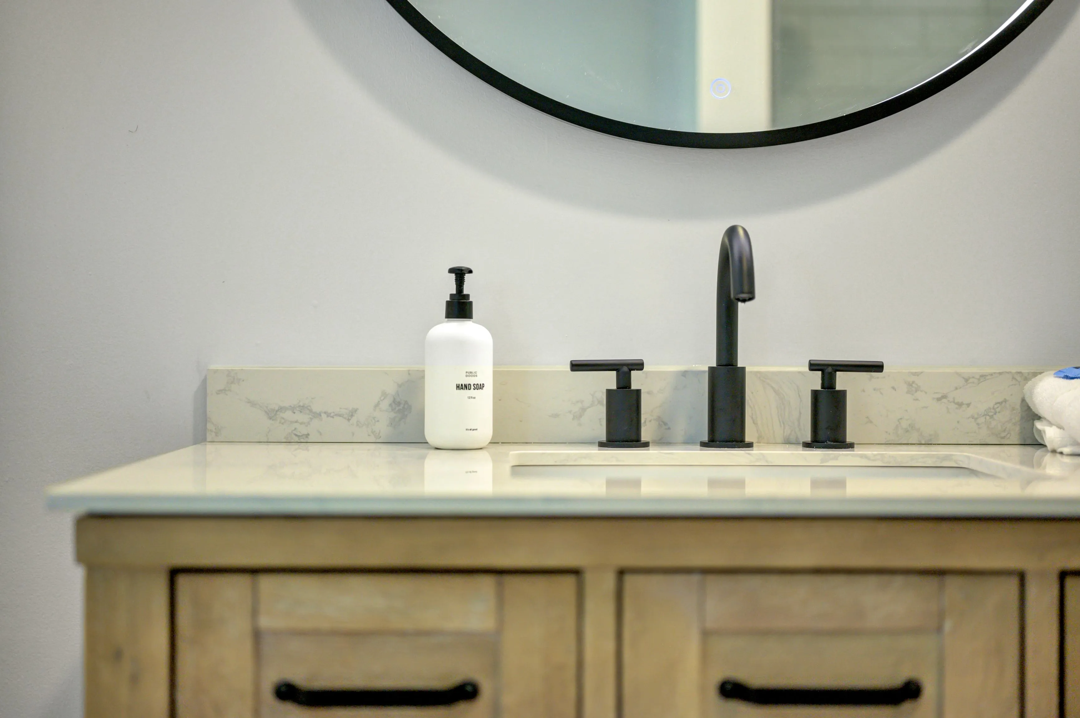 Modern bathroom sink with marble countertop, black faucet, and hand soap dispenser, reflected in a round mirror on a light-coloured wall.