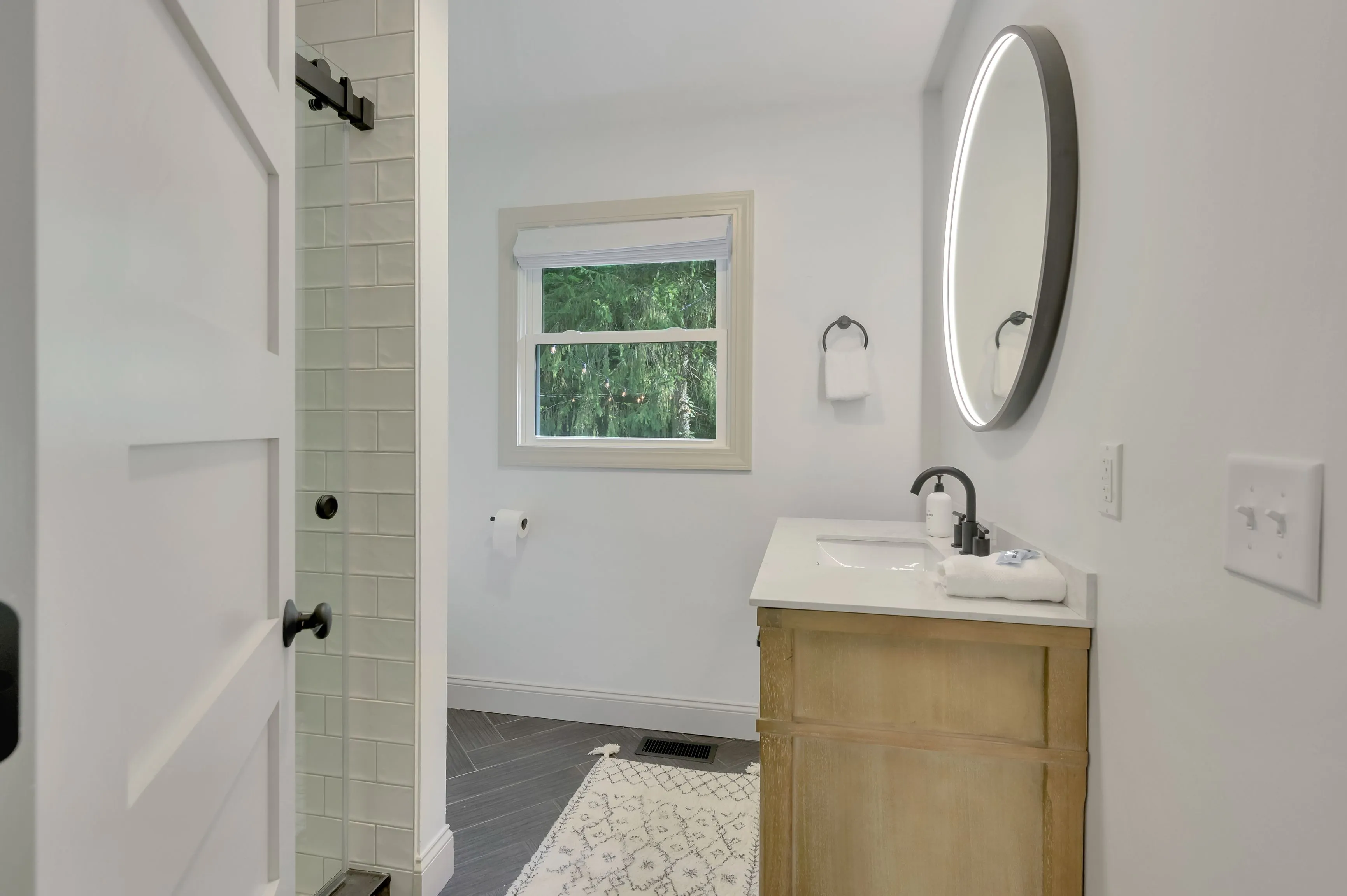Modern bathroom interior with a wooden vanity, white sink, large round mirror, a window with greenery outside, white subway tiles in the shower, and a patterned rug on the floor.