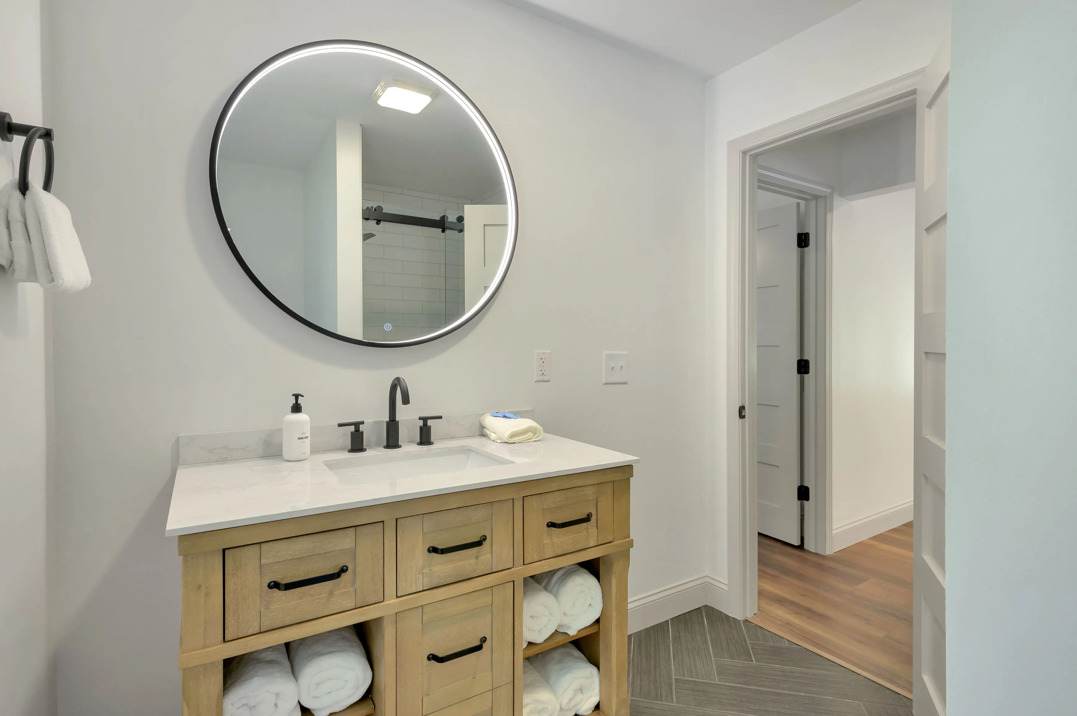 Modern bathroom interior with round mirror, wooden vanity with white countertop, and a view into the adjacent room.