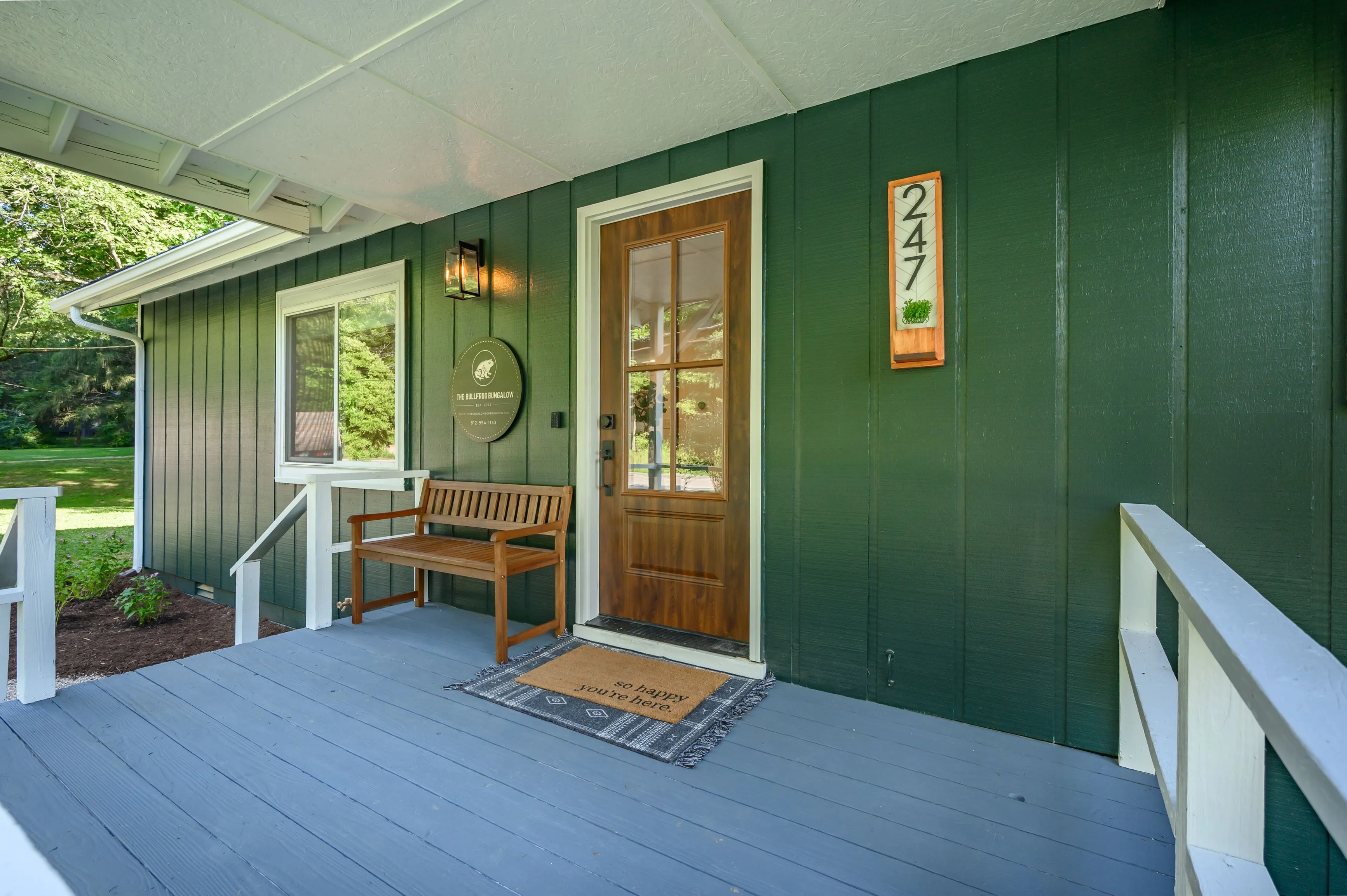 Home entrance with a dark green exterior, wooden front door with glass panes, a welcoming doormat, a bench, house number 247, and decorative elements.