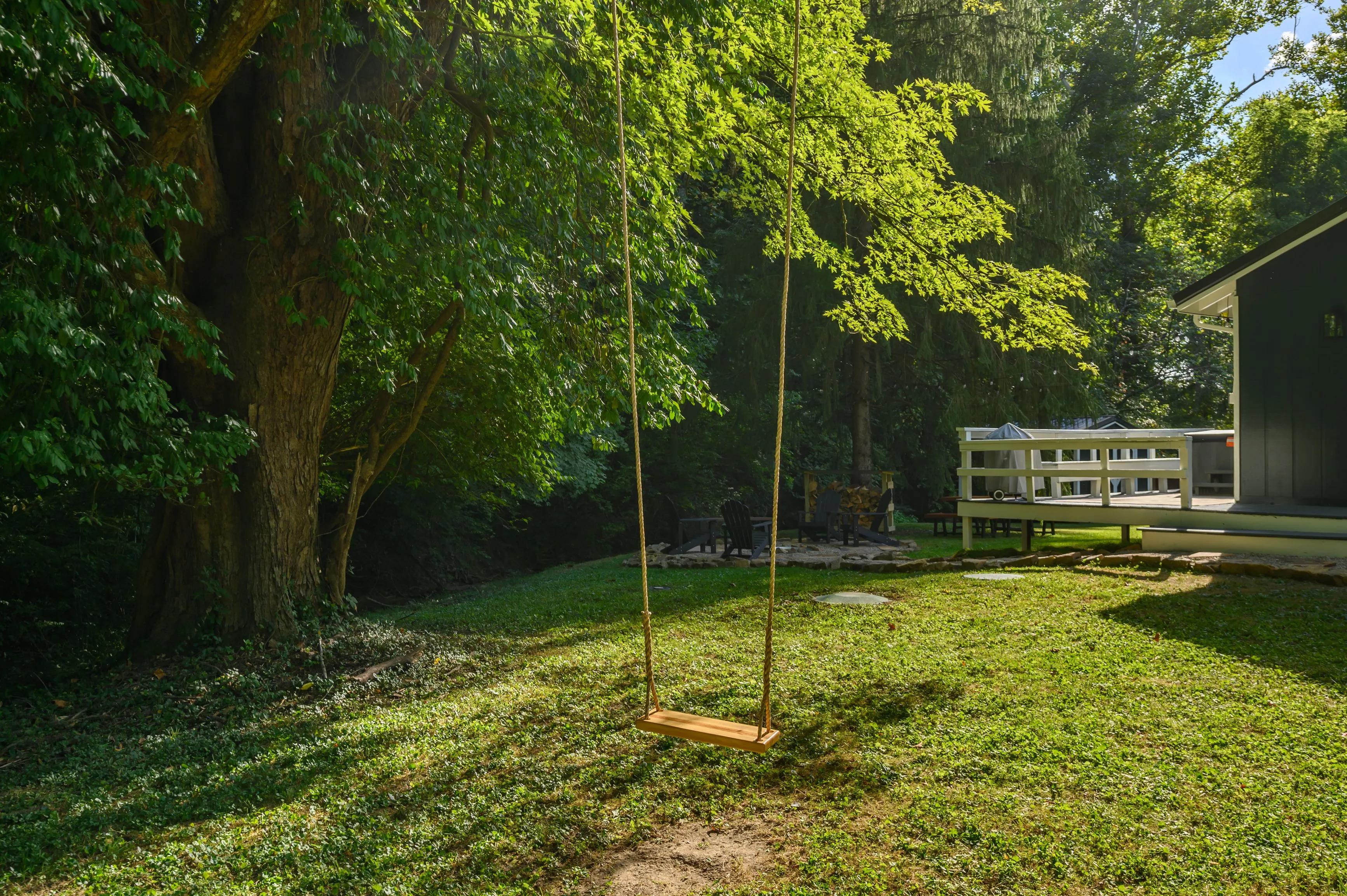 A wooden swing hanging from a tree in a lush green backyard with a playhouse in the background and sunlight filtering through the leaves.