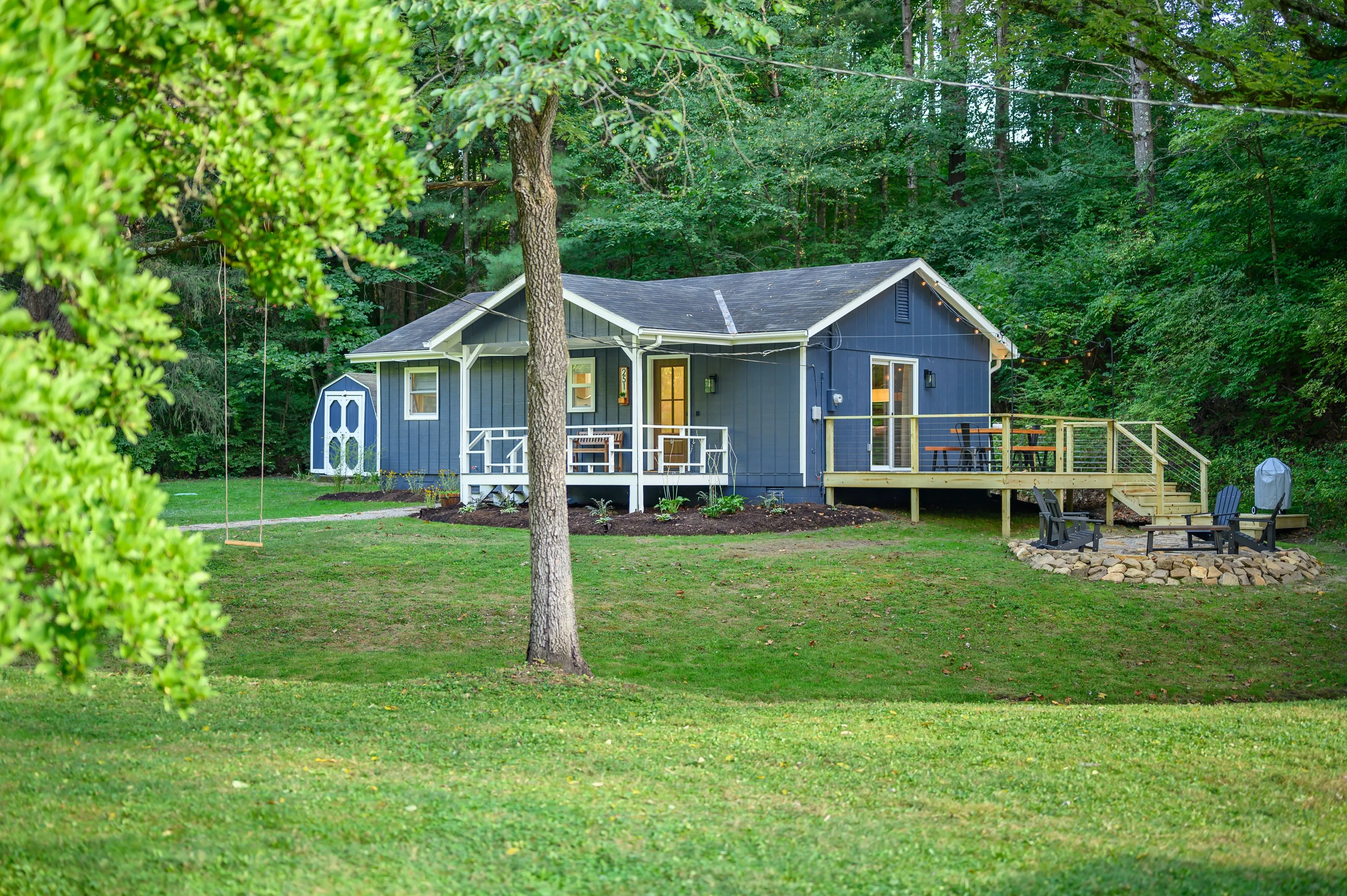 A cozy grey house with white trim and a front porch, nestled in a green lawn with surrounding trees, featuring a wooden swing and a fire pit area with chairs.