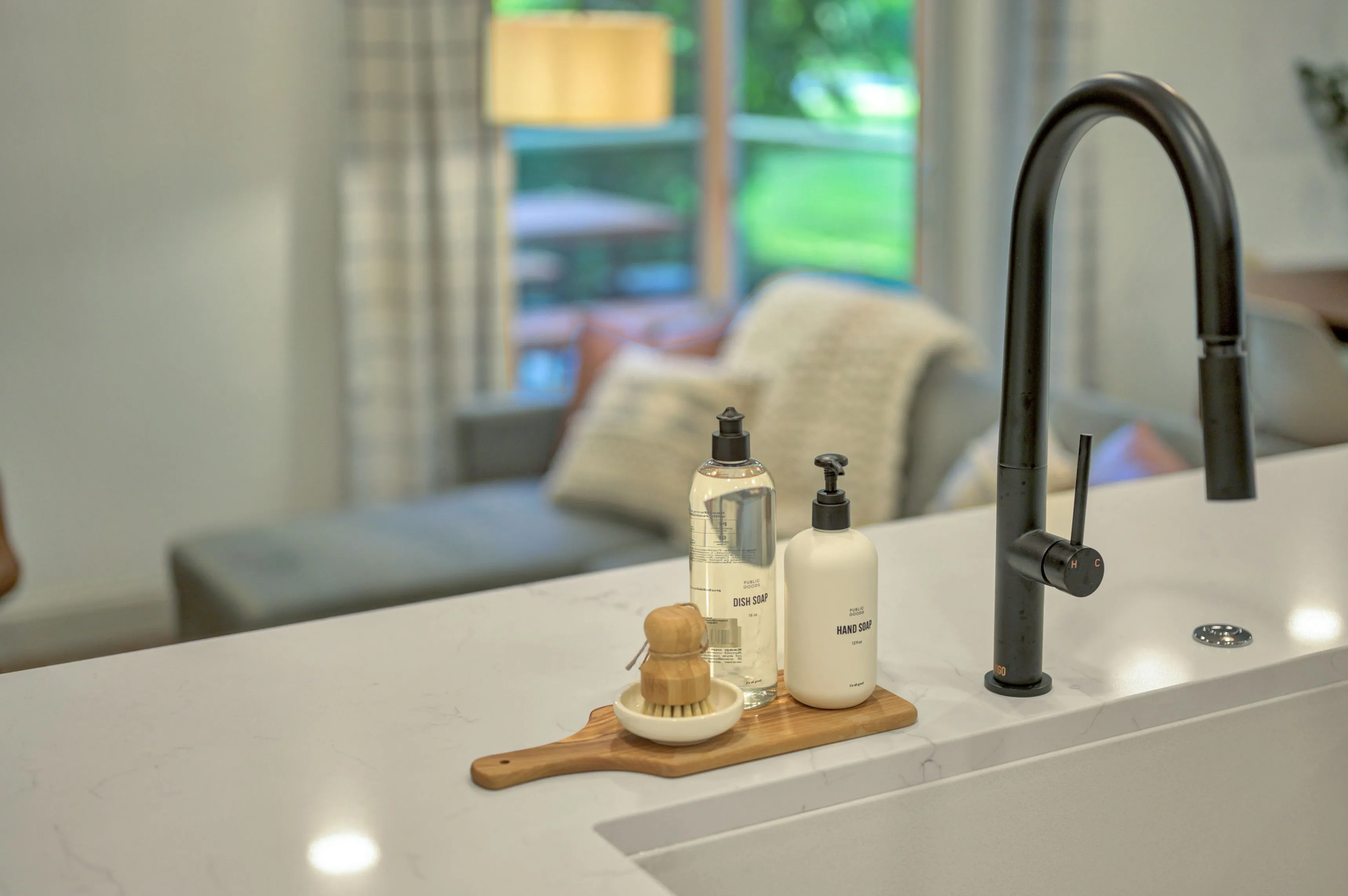 Modern kitchen sink with a black faucet, soap dispensers, and a wooden dish brush on a marble countertop, with a blurred living room background.