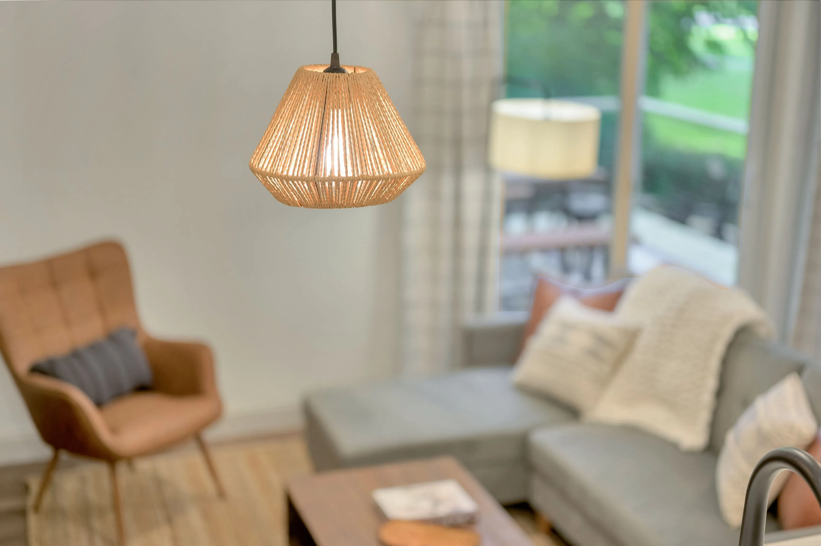 A warmly lit interior with a focus on a stylish pendant lamp hanging above a wooden coffee table, with a blurred background showcasing a cozy living room corner with an armchair and a couch adorned with cushions.