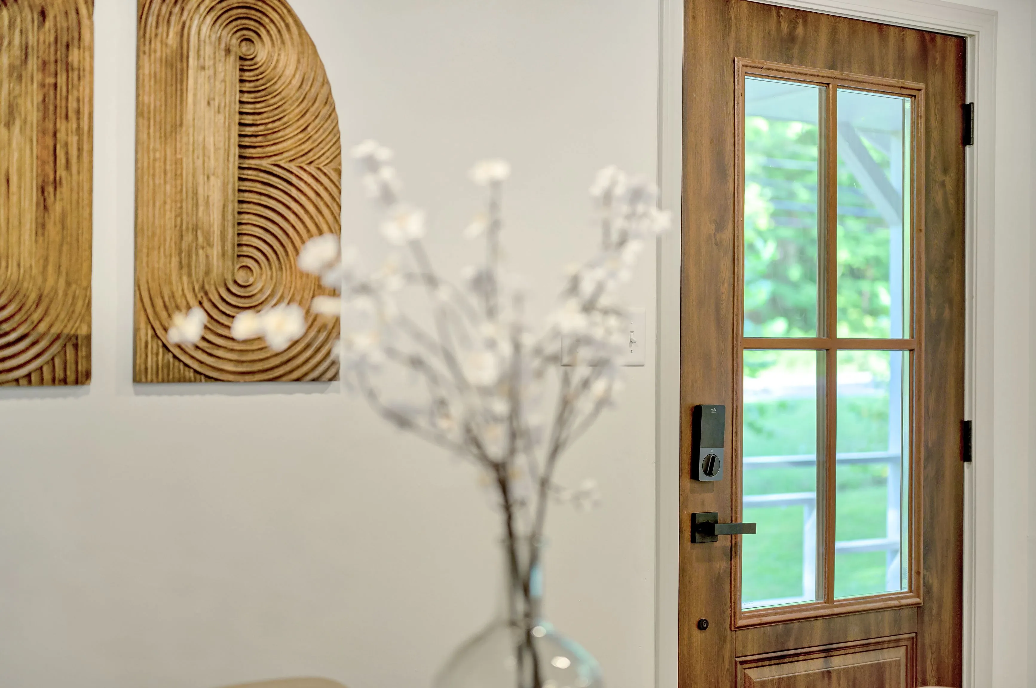 A modern interior hallway featuring a wooden door with glass panes, two abstract wooden wall art pieces, and a blurred foreground of a glass vase with white branches.