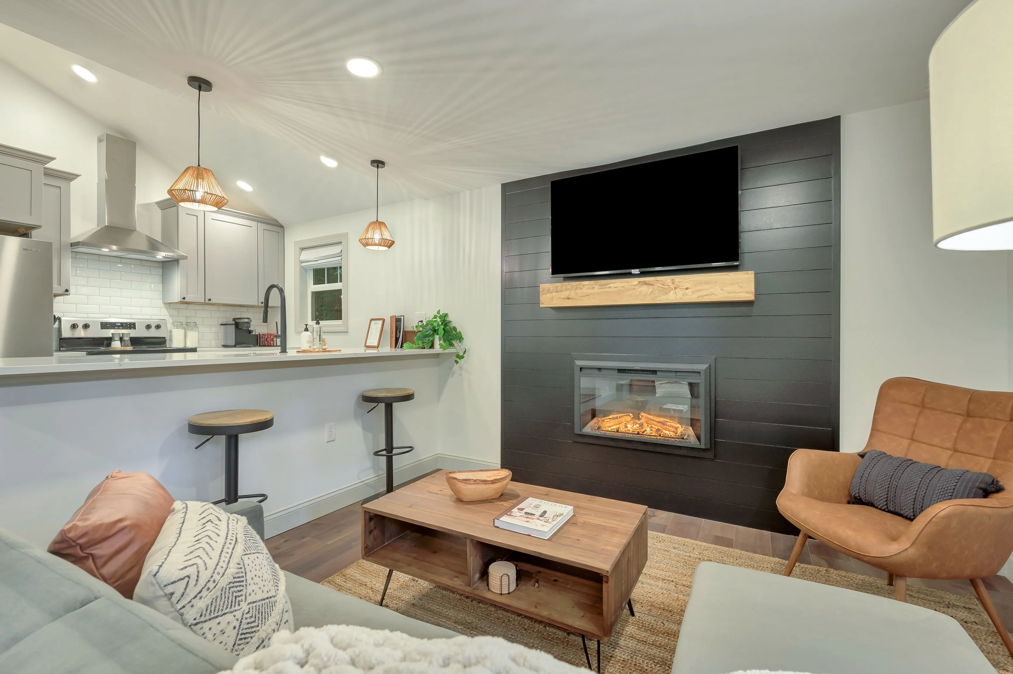 Modern living room seamlessly connected to a kitchen featuring a fireplace with a mounted TV above, cozy furnishings, and contemporary lighting.