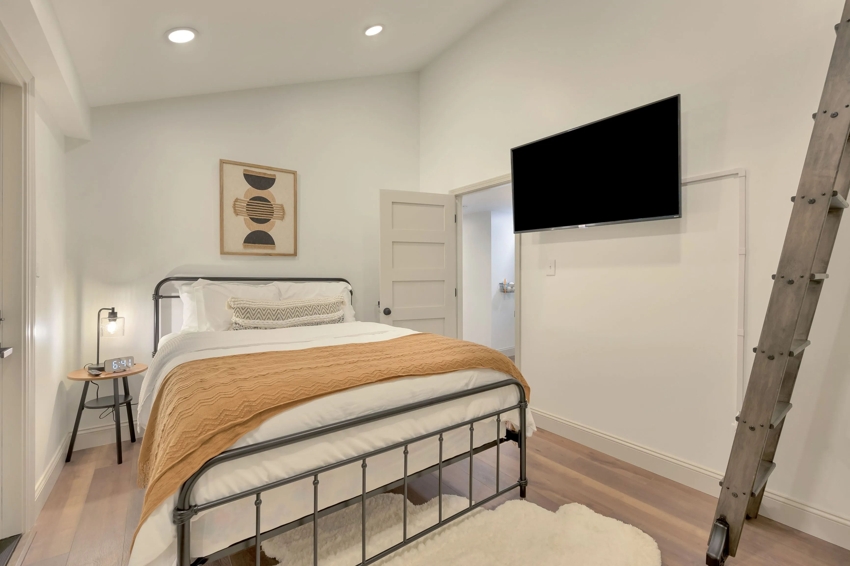 Modern bedroom interior with a metal frame bed, white and patterned bedding, a mounted flat-screen TV, and decorative artwork above the bed. A rustic wooden ladder rests against the wall on the right, with soft lighting and a neutral color palette.