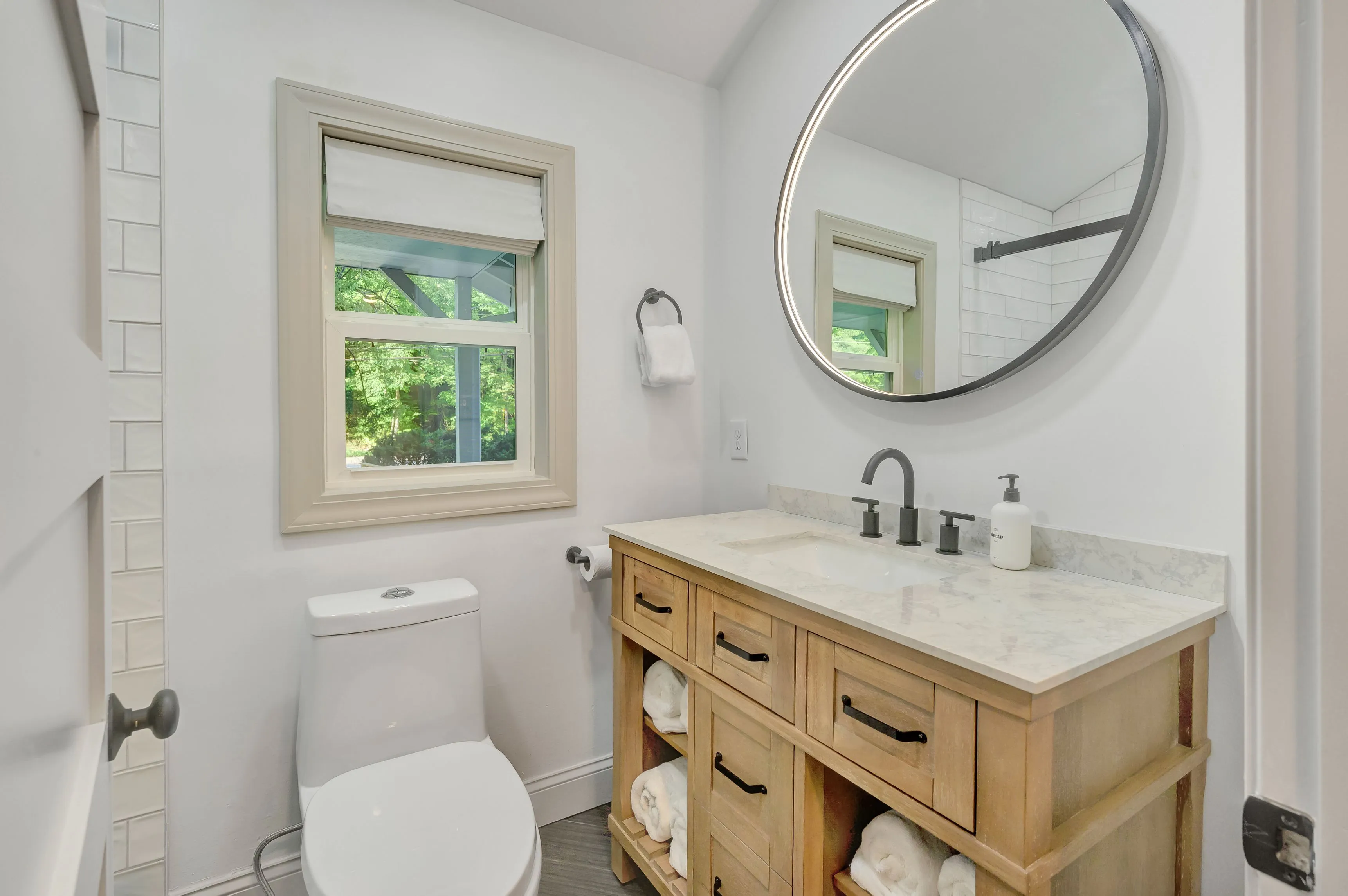 Modern bathroom interior with wooden vanity cabinet, marble countertop, round mirror, and view of greenery outside the window.