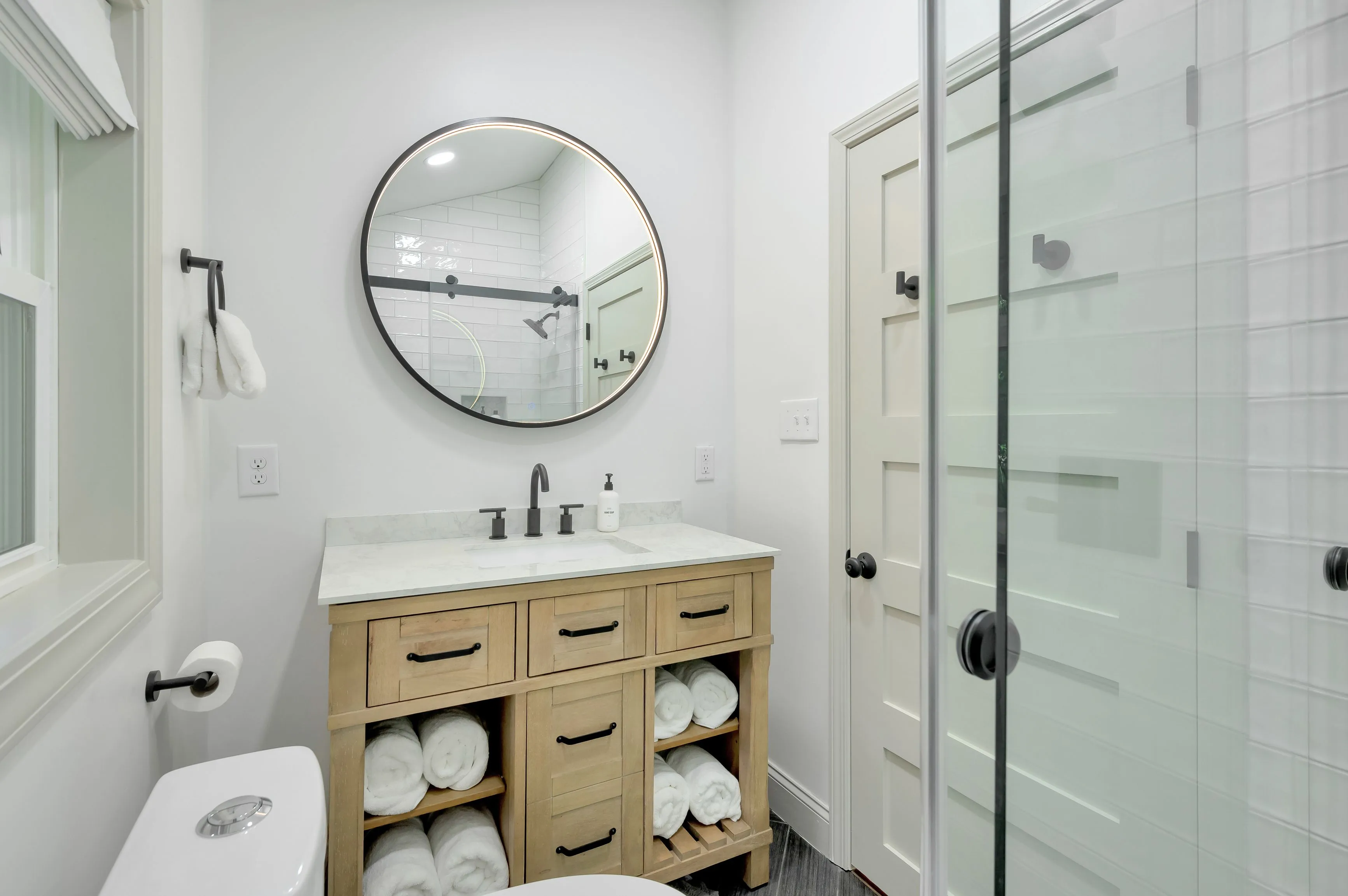 Modern bathroom interior with round mirror, wooden vanity cabinet, white towels, and glass shower enclosure.