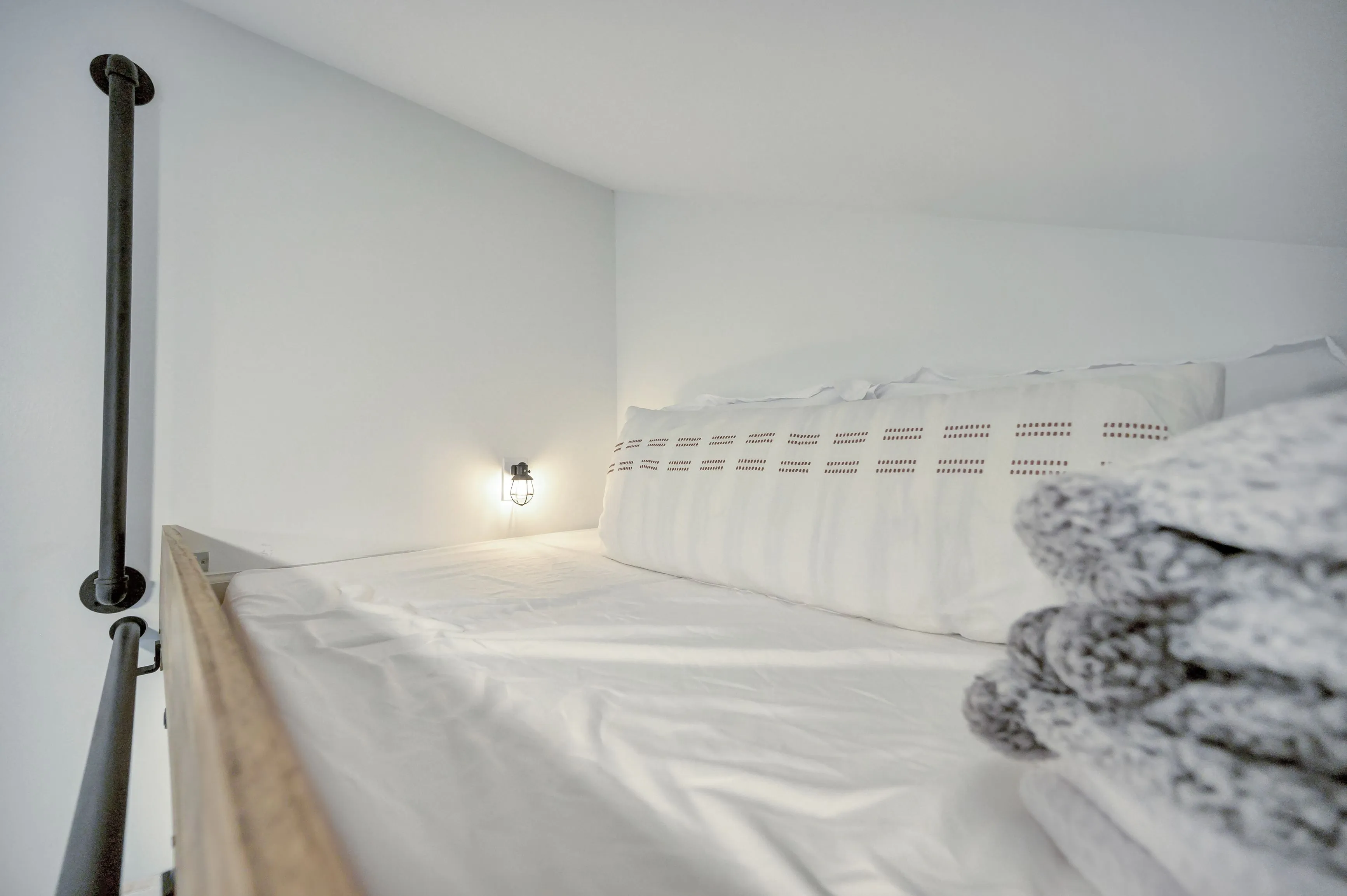 Minimalist bedroom with a white bed, pillows, and a gray knitted blanket, featuring a wall-mounted light and a dark handrail.