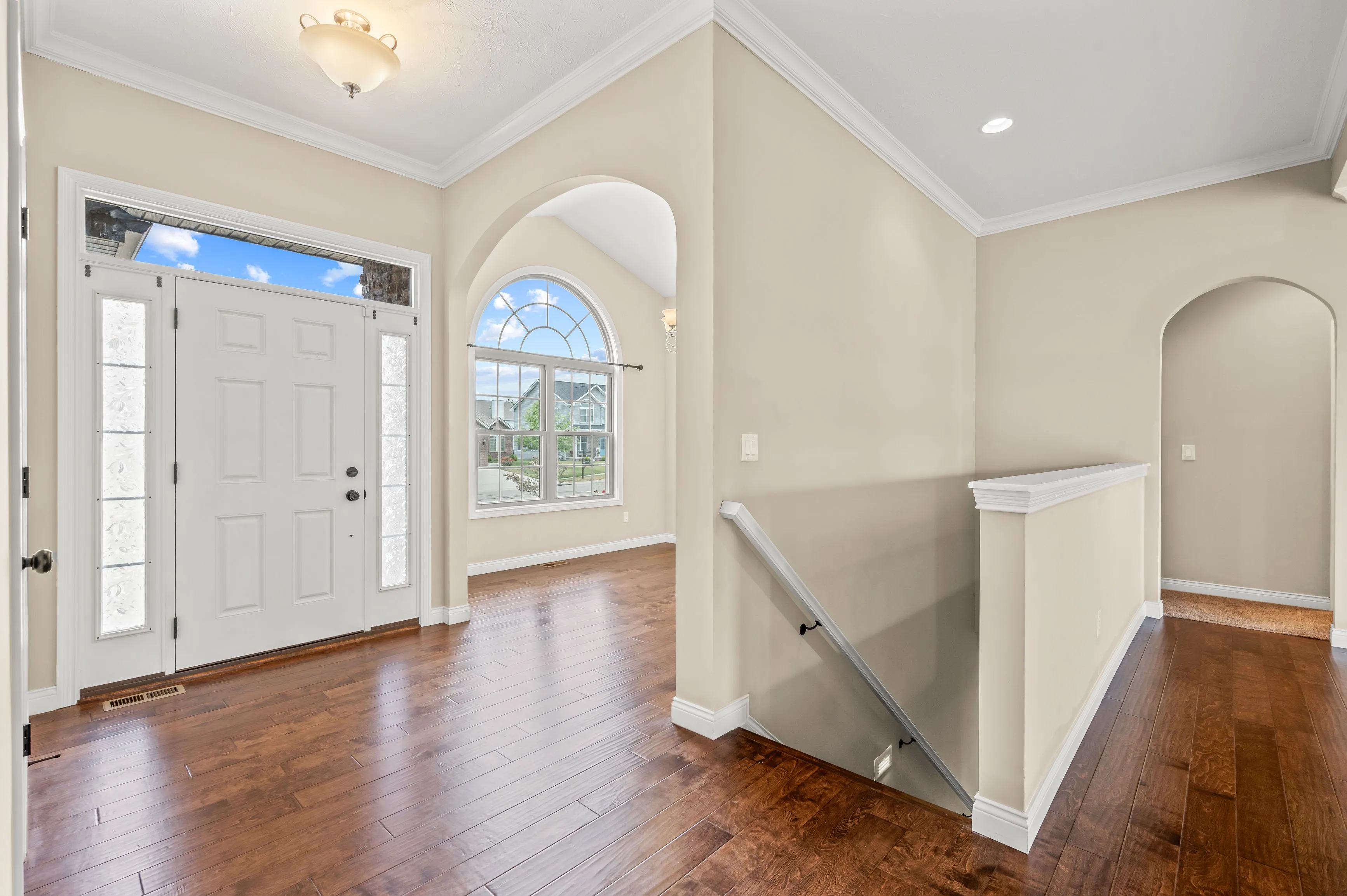 Empty living room with hardwood floors, white walls, a large arched window, and a front door with sidelights.