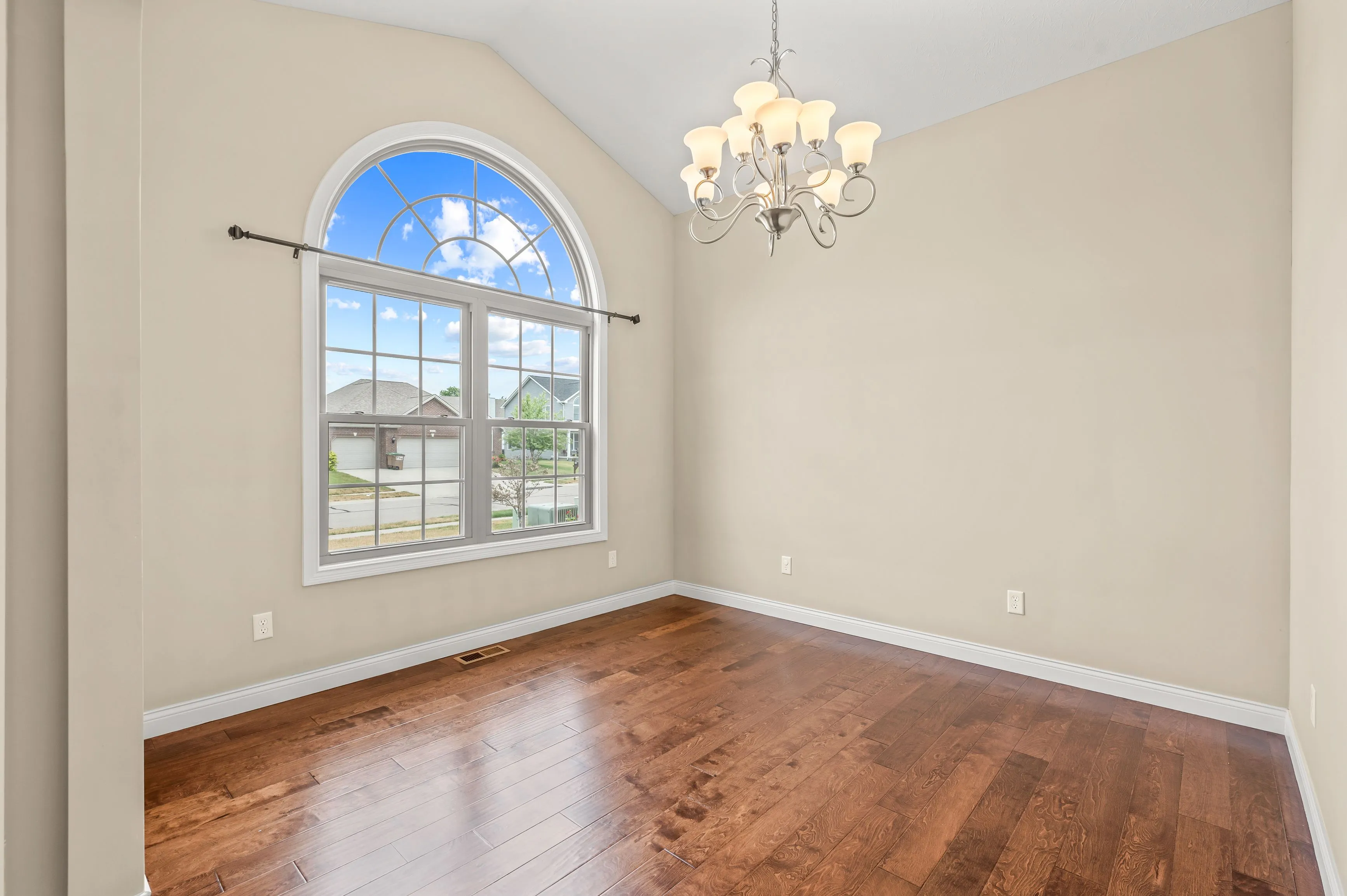 Empty room with polished hardwood floor, beige walls, a large arched window, and a chandelier.
