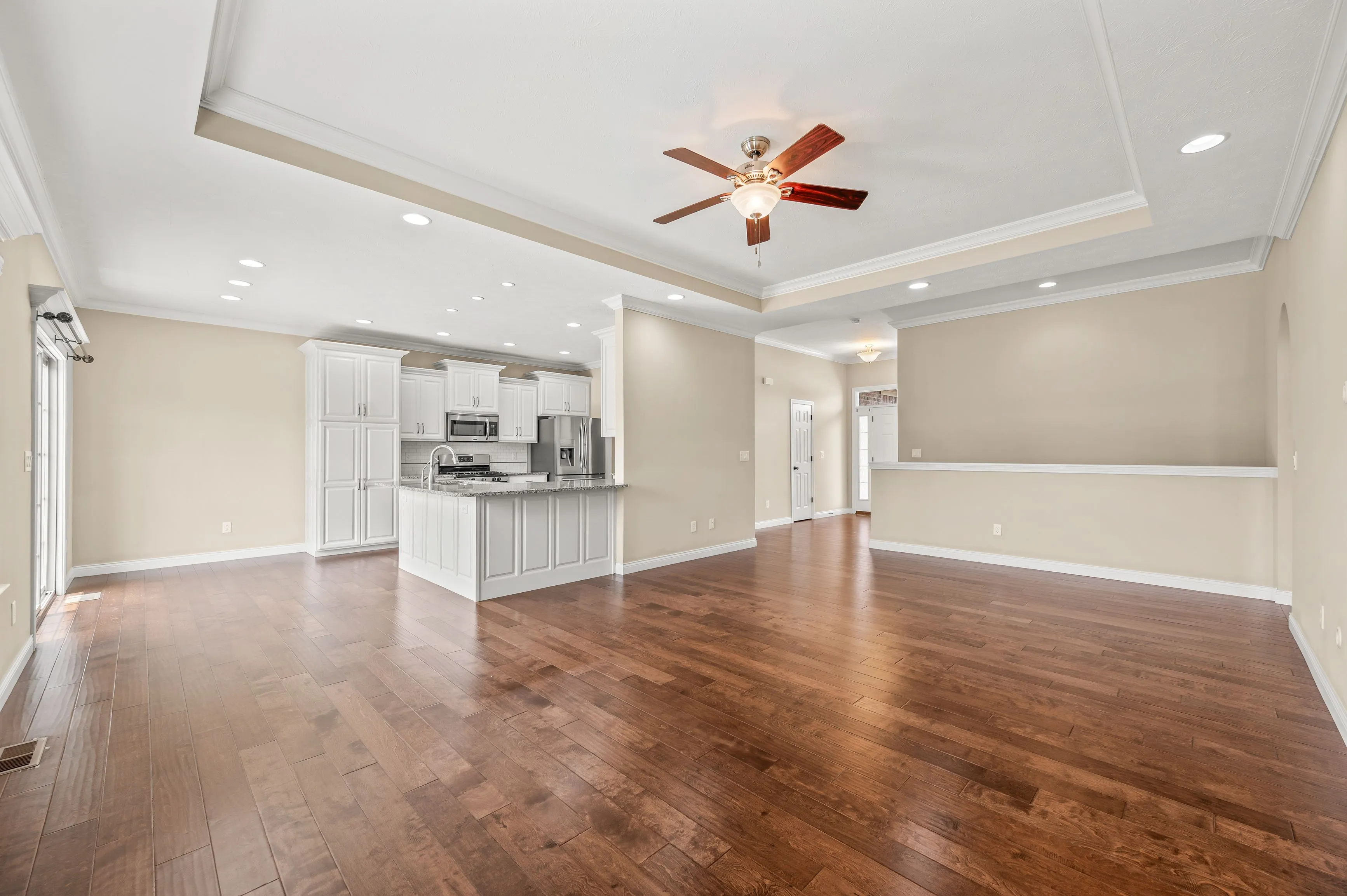 Empty spacious interior of a house with wooden floors, white walls, and a kitchen area in the background with a ceiling fan.