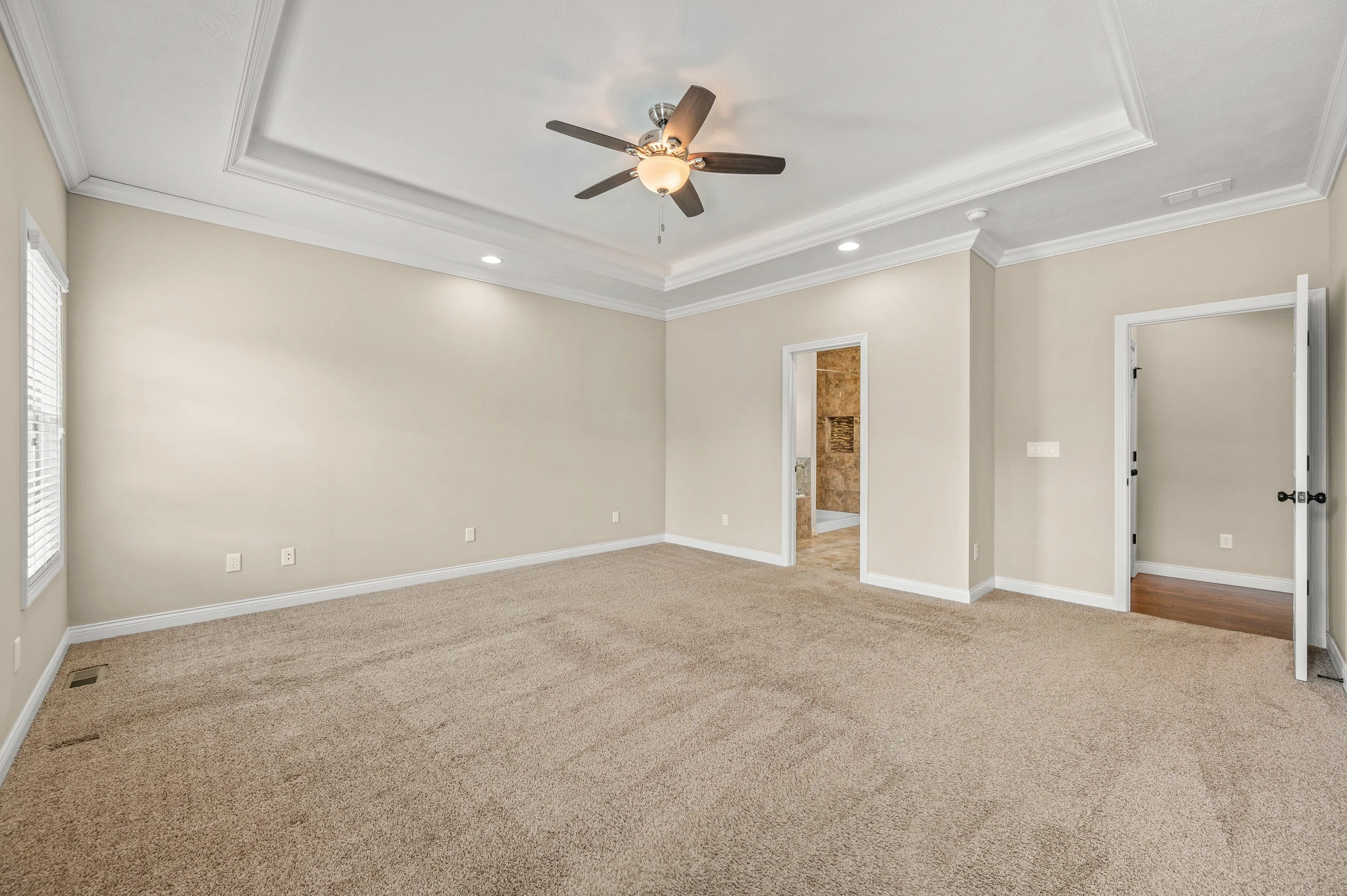 Empty room with beige walls, carpeted floor, ceiling fan, and open doors leading to adjacent rooms.