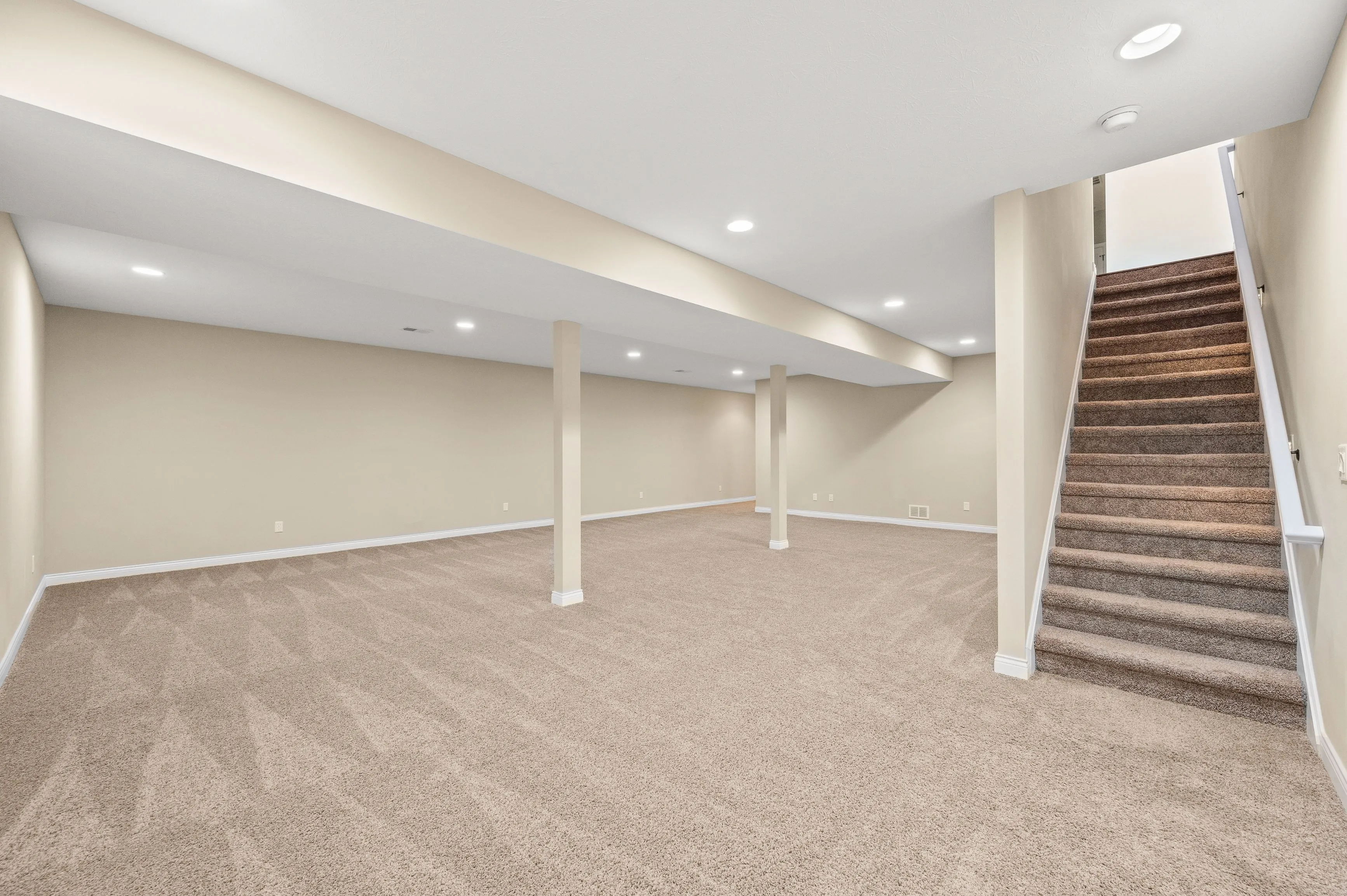 Empty basement interior with beige carpeting, white walls with several support columns, and a staircase to the upper level.