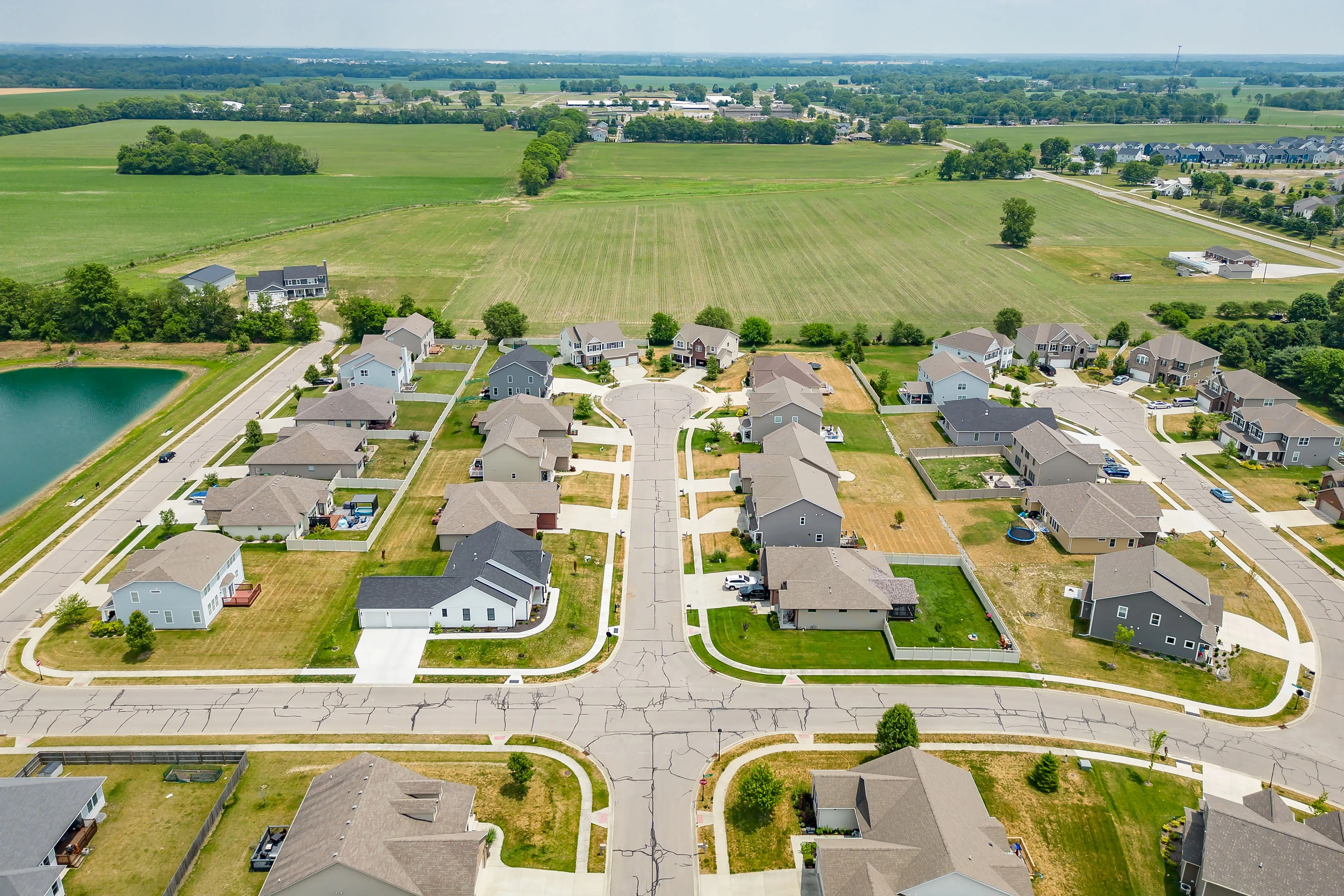 Aerial view of a suburban neighborhood with rows of houses, manicured lawns, and a small lake nearby.
