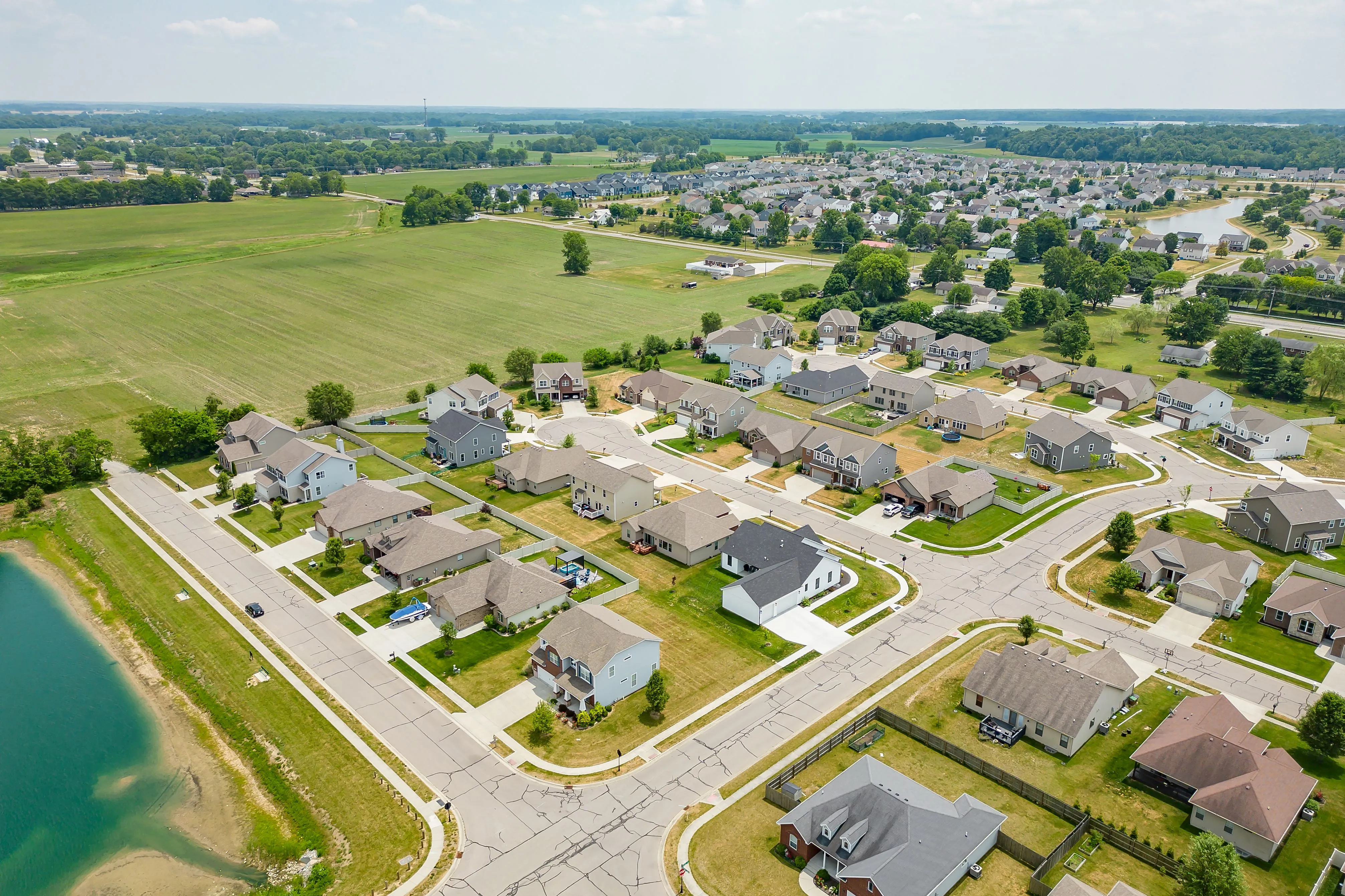 Aerial view of a suburban neighborhood next to a body of water with green open fields in the background.