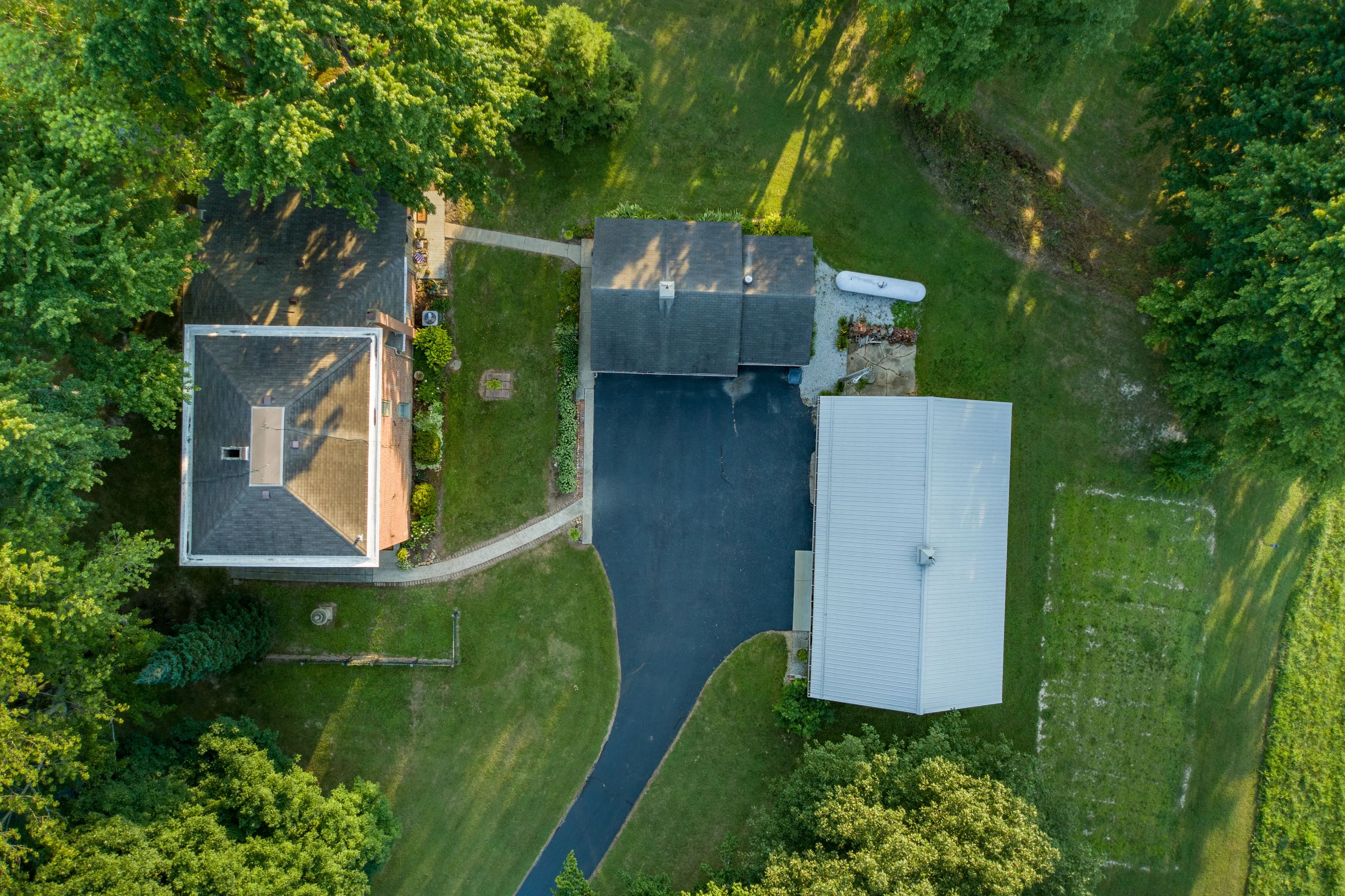 Aerial view of two houses with dark roofs surrounded by green trees and lawns with a curved driveway connecting them.