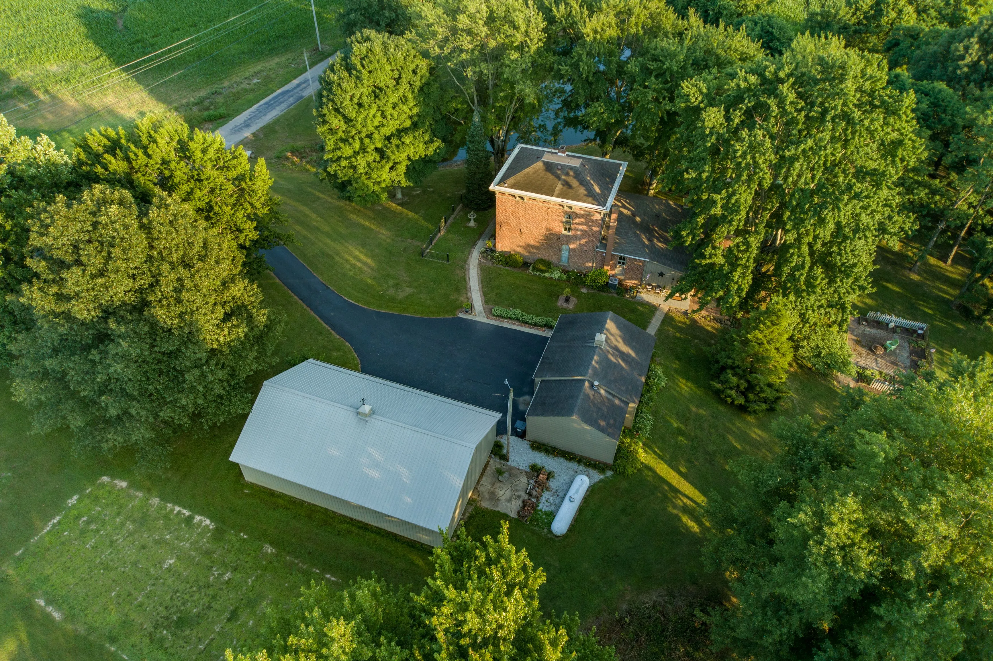 Aerial view of a residential property with a brick house, large metal outbuilding, and surrounding green trees.