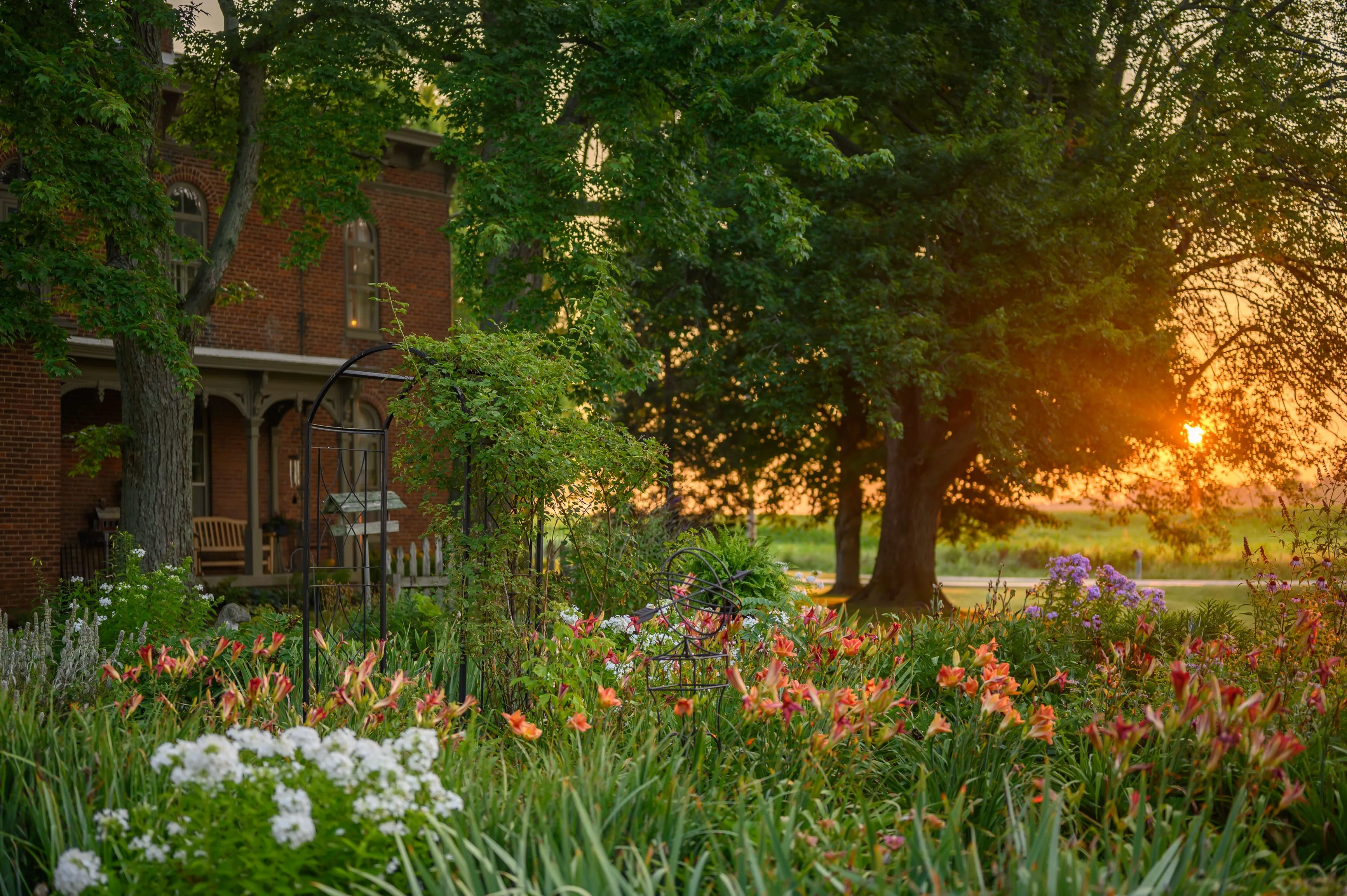 Idyllic brick country house with porch, surrounded by lush garden and trees, bathed in warm sunlight at sunset.