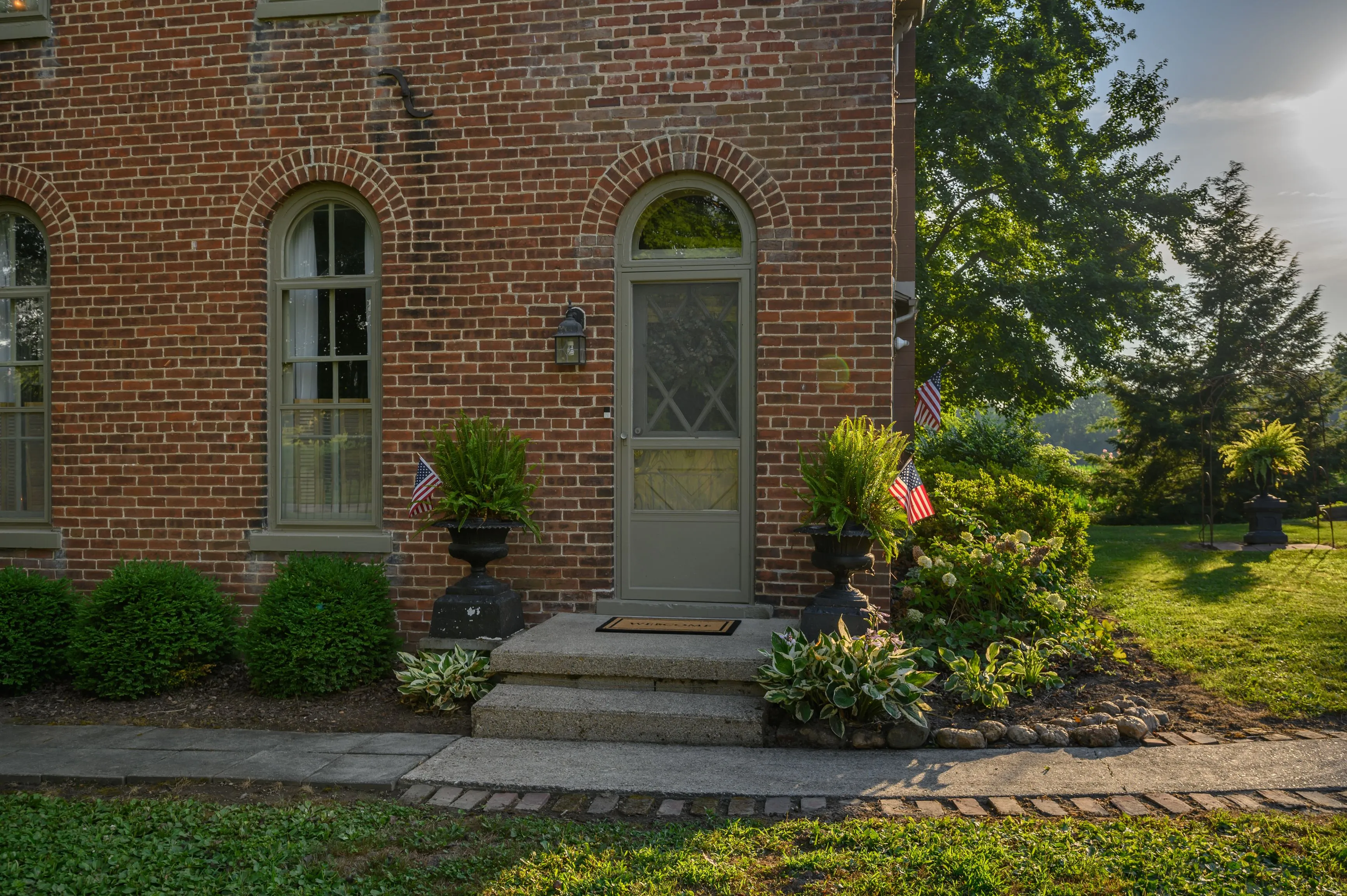 Brick house entrance with arched doorway, stone steps, adorned by potted plants and American flags, during early morning sunlight.