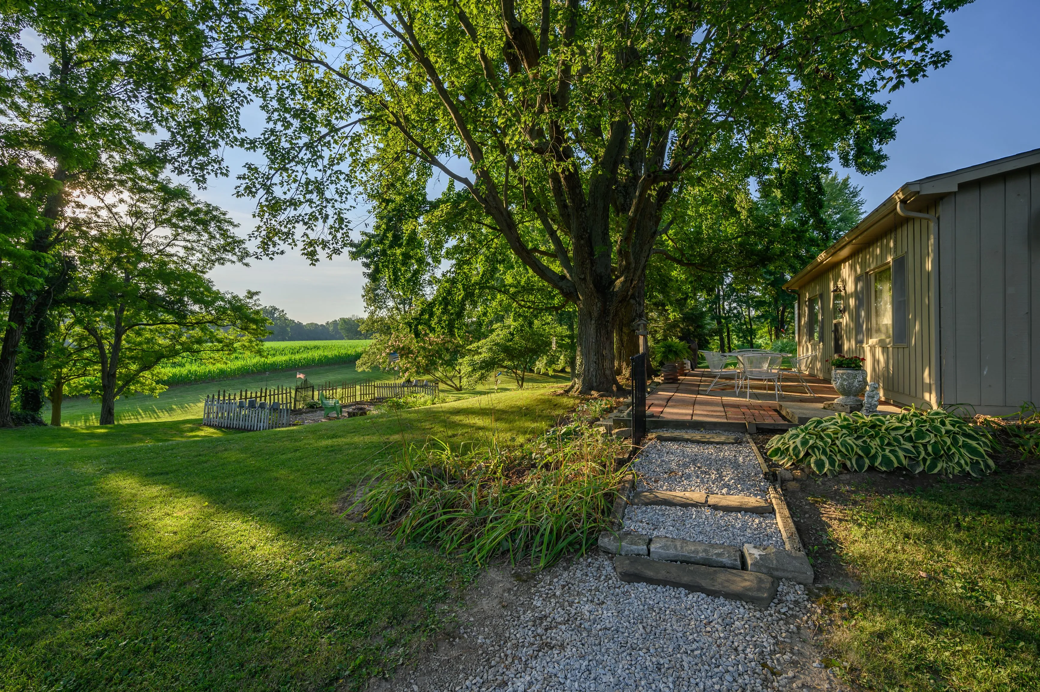 A serene backyard with a large tree, gravel path, patio furniture, and a view of green fields in the background.
