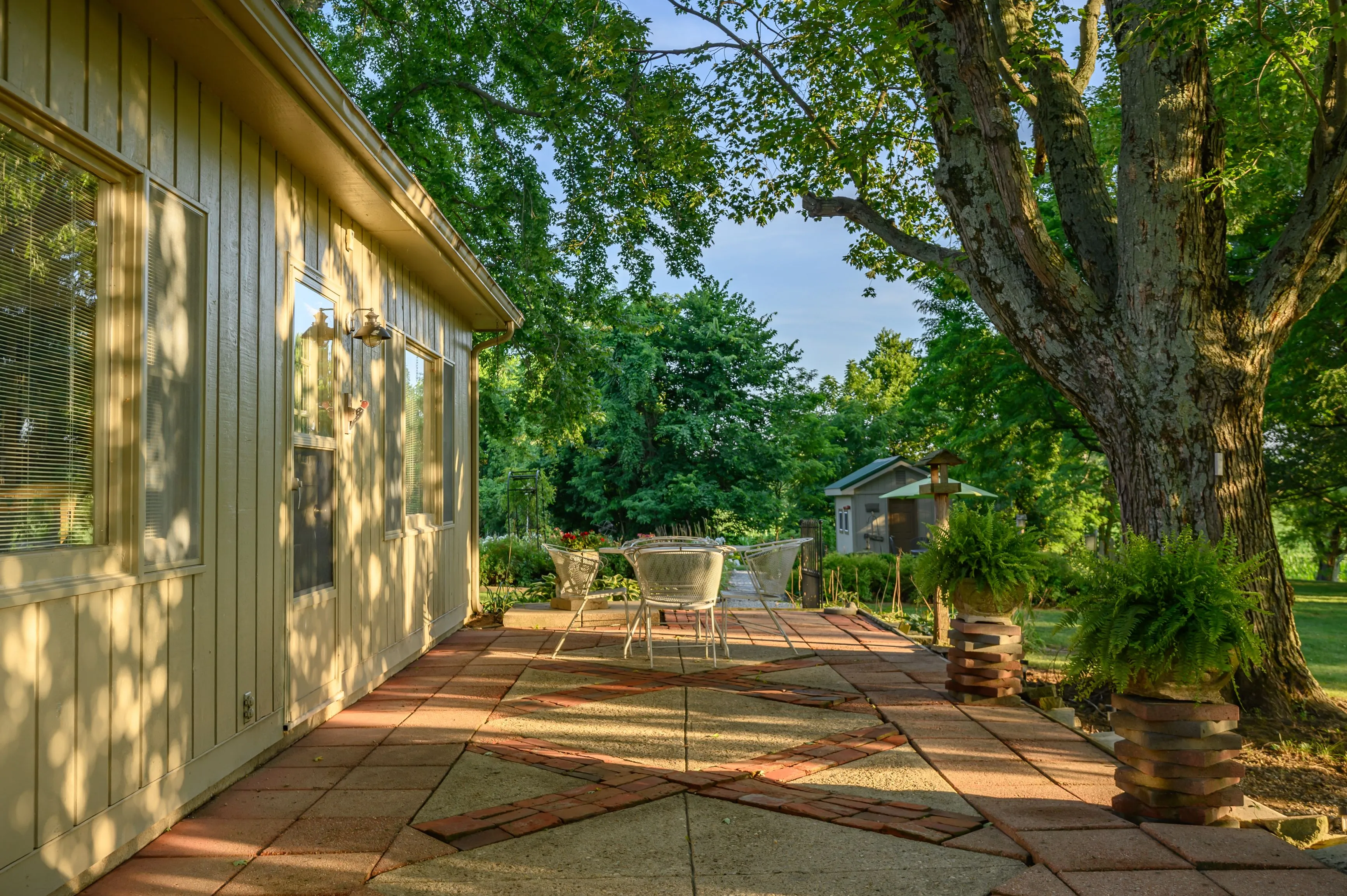 Patio with brick flooring, outdoor furniture, and large shade tree beside a house during the day.