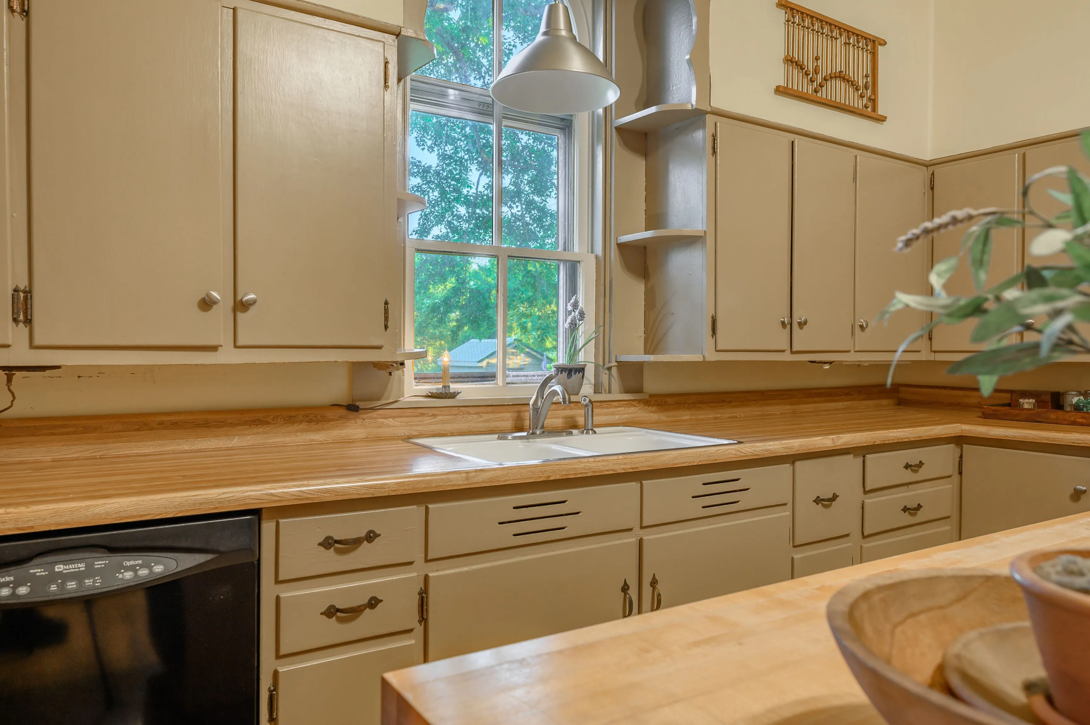 Cozy kitchen interior with wooden countertops, beige cabinets, a sink with a window above it, and greenery on the side.
