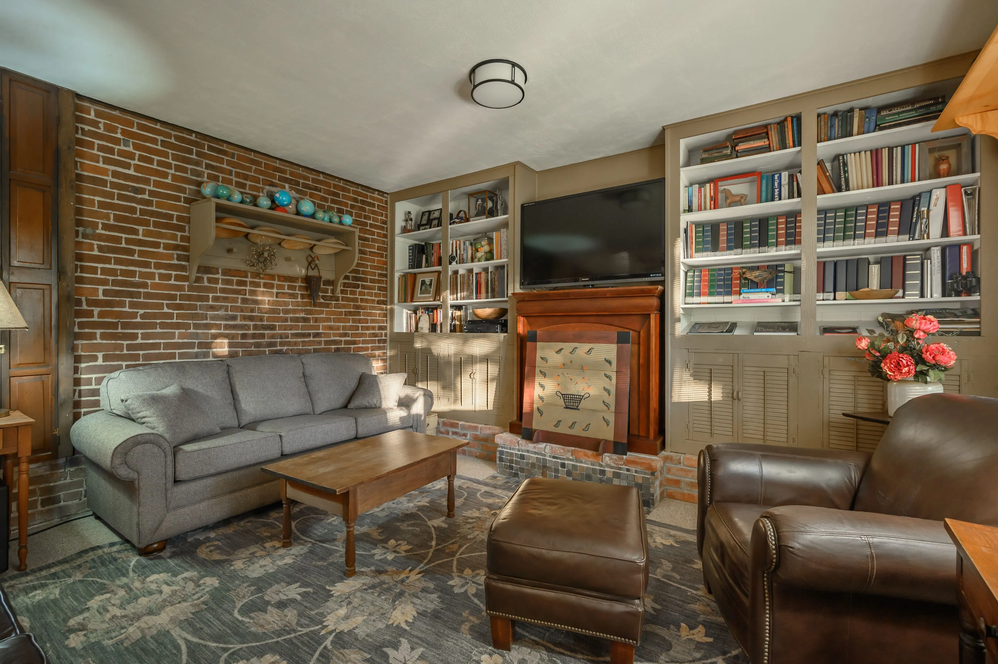 Cozy living room interior featuring a gray couch, leather armchair, wooden coffee table, bookshelves filled with books, a brick fireplace, and decorative elements.