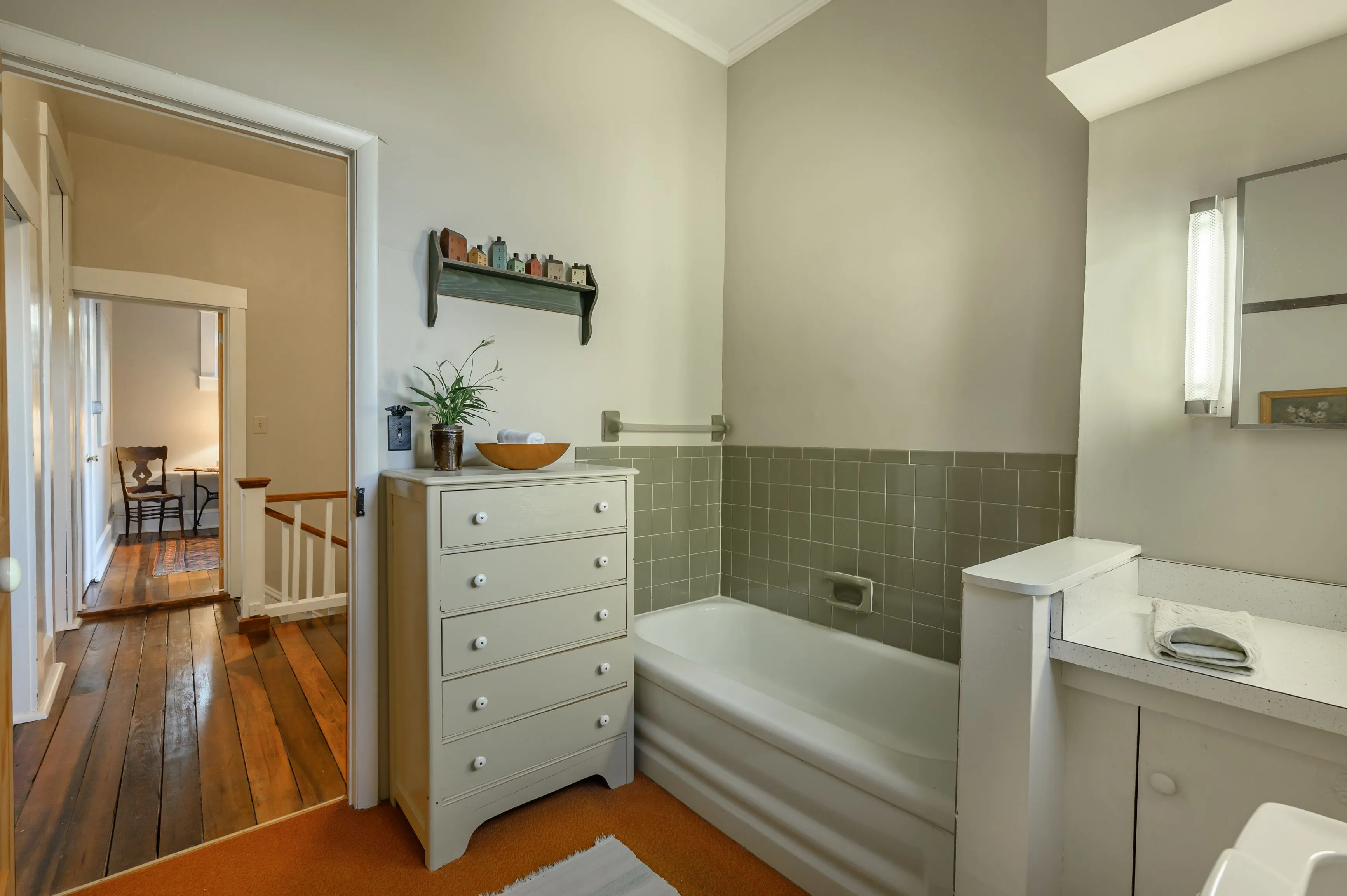 A cozy bathroom with a white bathtub and vanity, wooden floors, gray tiles, and a decorative shelf, with a view into a hallway.