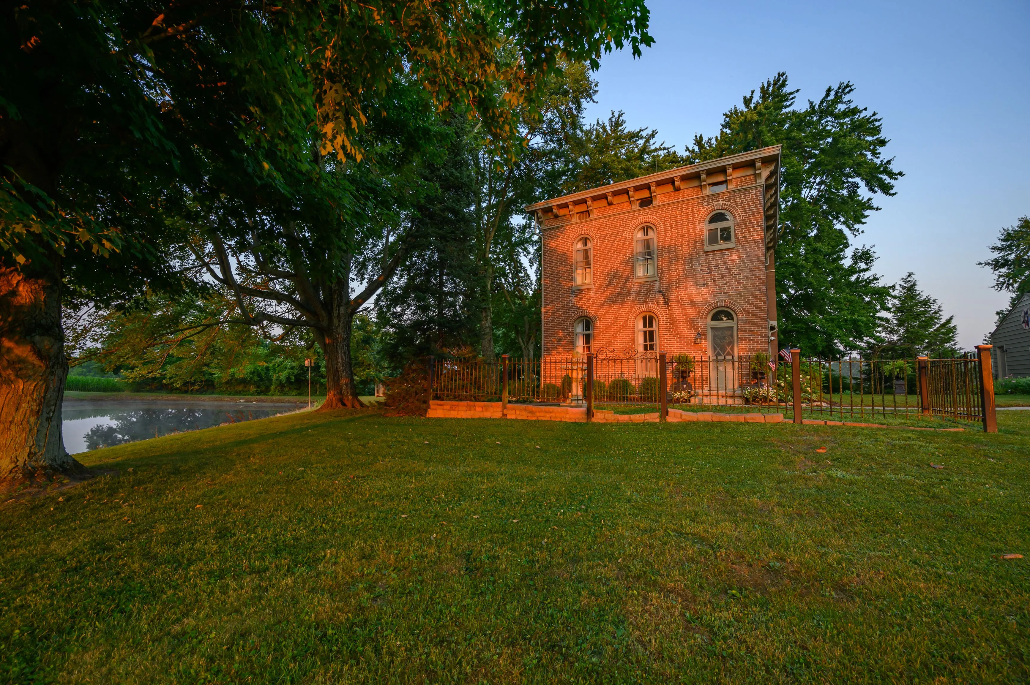 Brick building with wrought iron fence at sunset, surrounded by green lawn and trees, with a pond in the background.