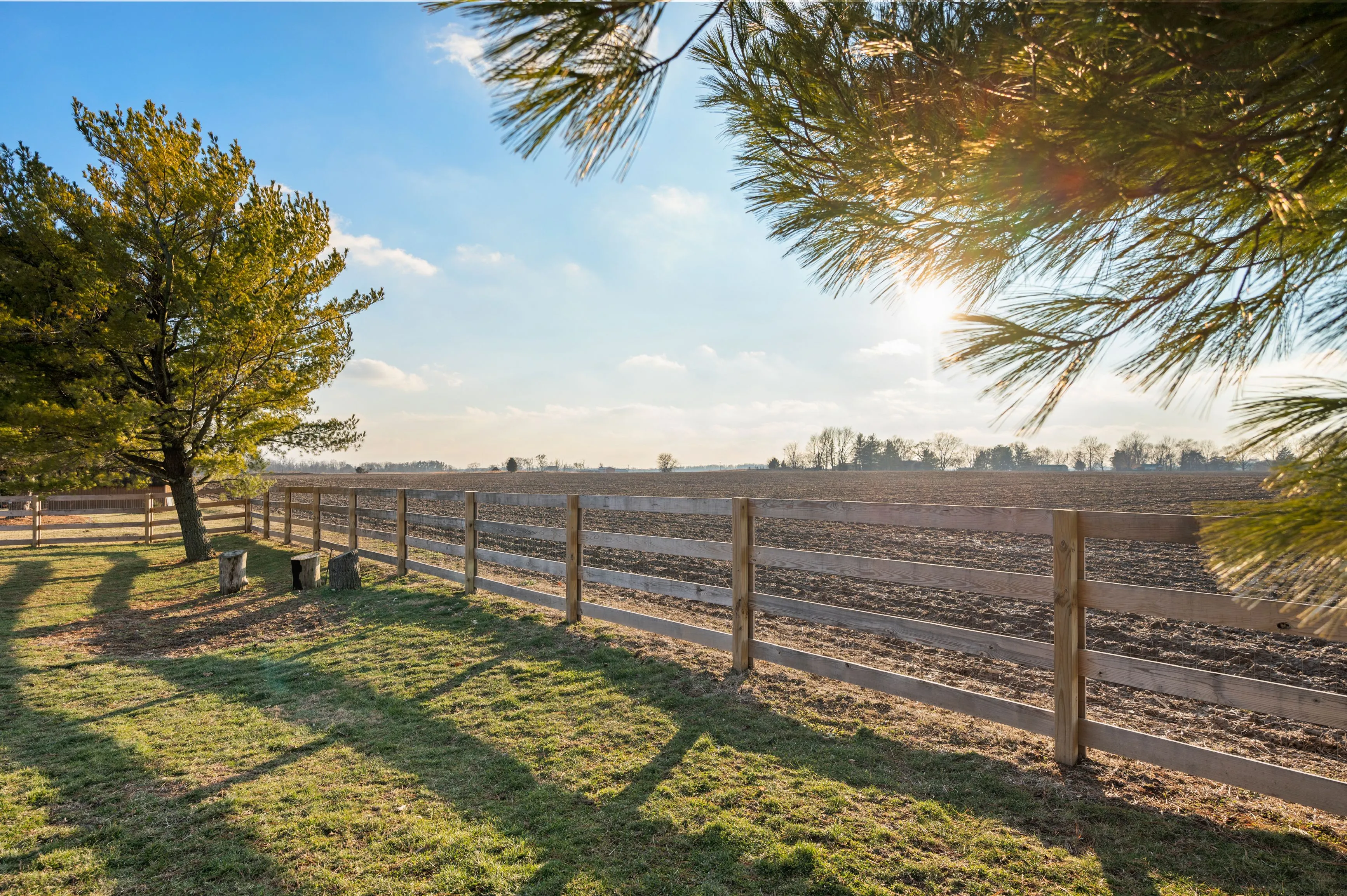 Rural landscape with a wooden fence, trees, and plowed field under a clear sky with sunbeams.