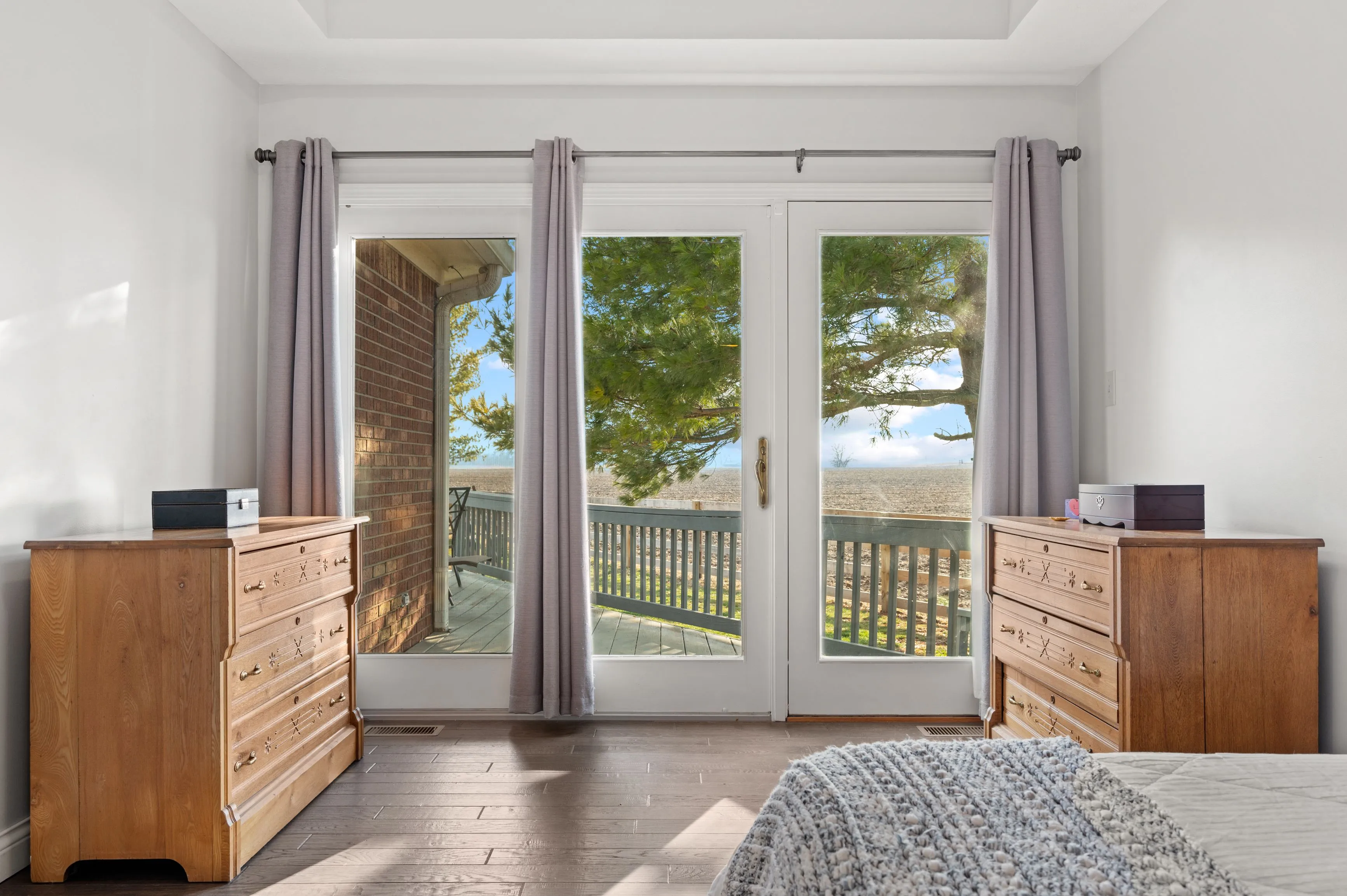 Bright bedroom with open French doors leading to a balcony with a view of trees and the horizon, flanked by gray curtains and furnished with a wooden dresser and a glimpse of a covered bed in the foreground.