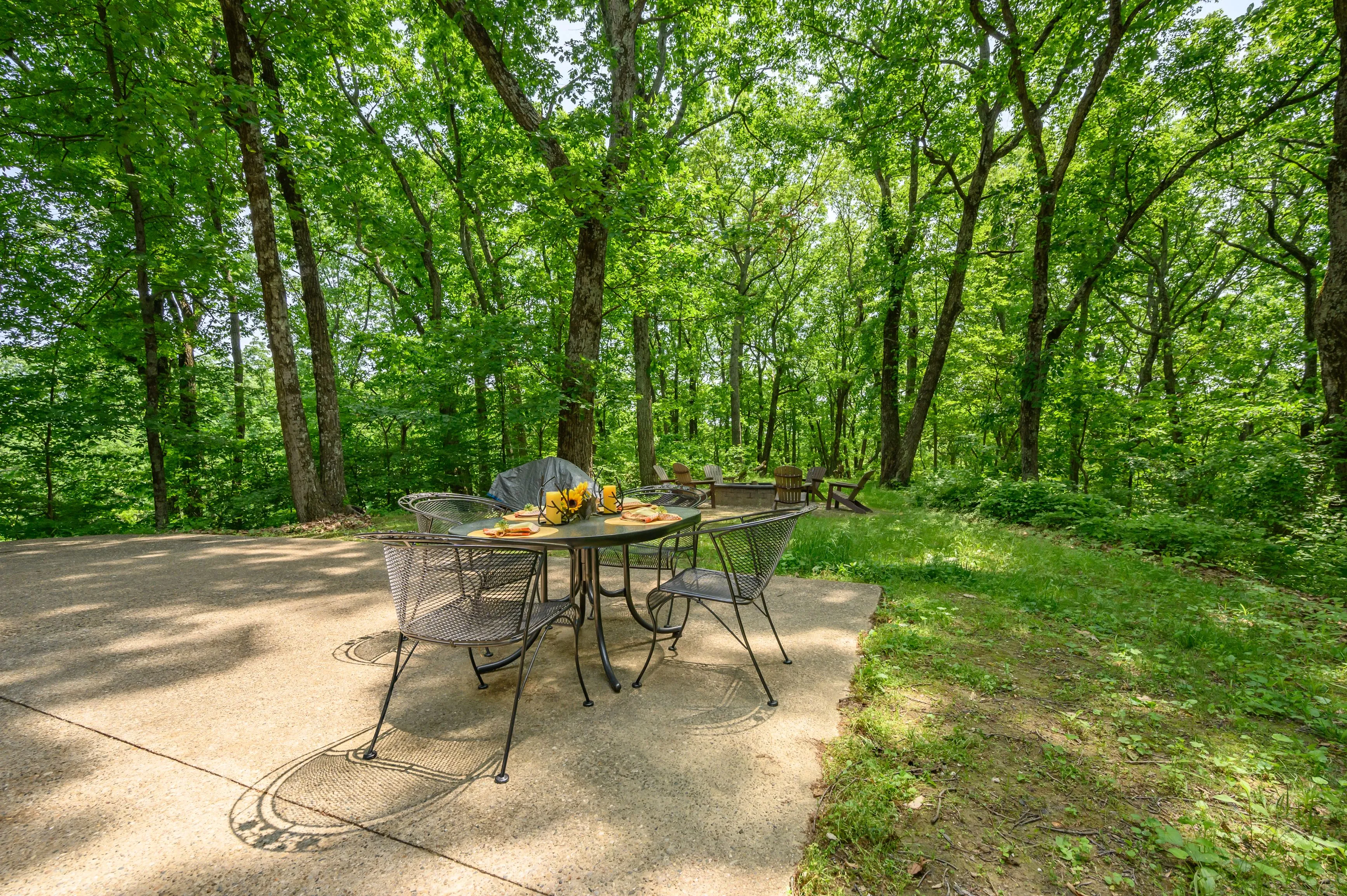 Patio dining set on a concrete slab in the woods with a table set with food and drinks, surrounded by lush green trees.