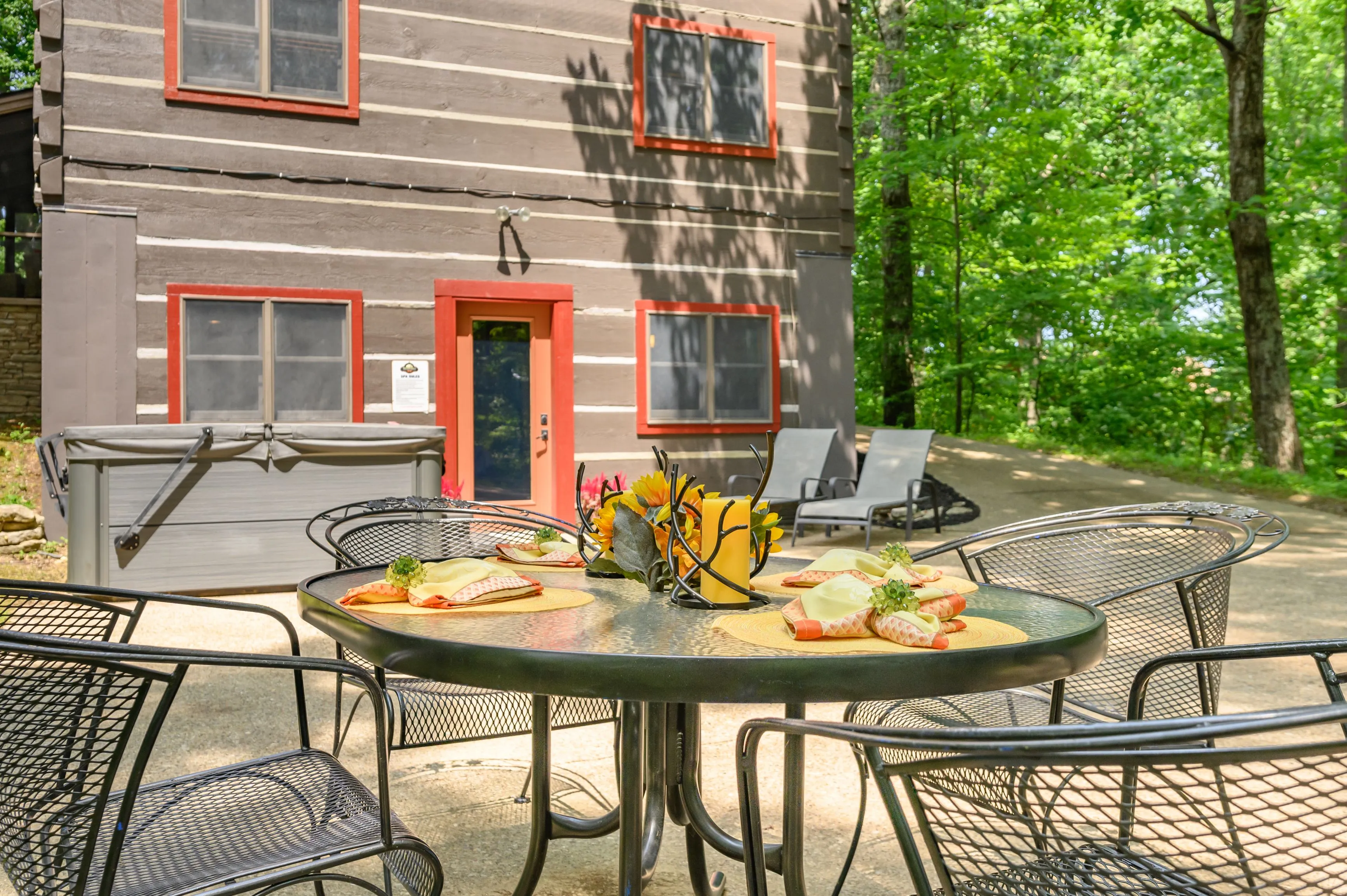 Outdoor patio setting with a meal prepared on a table, in front of a two-story house with red accents and surrounded by green trees.