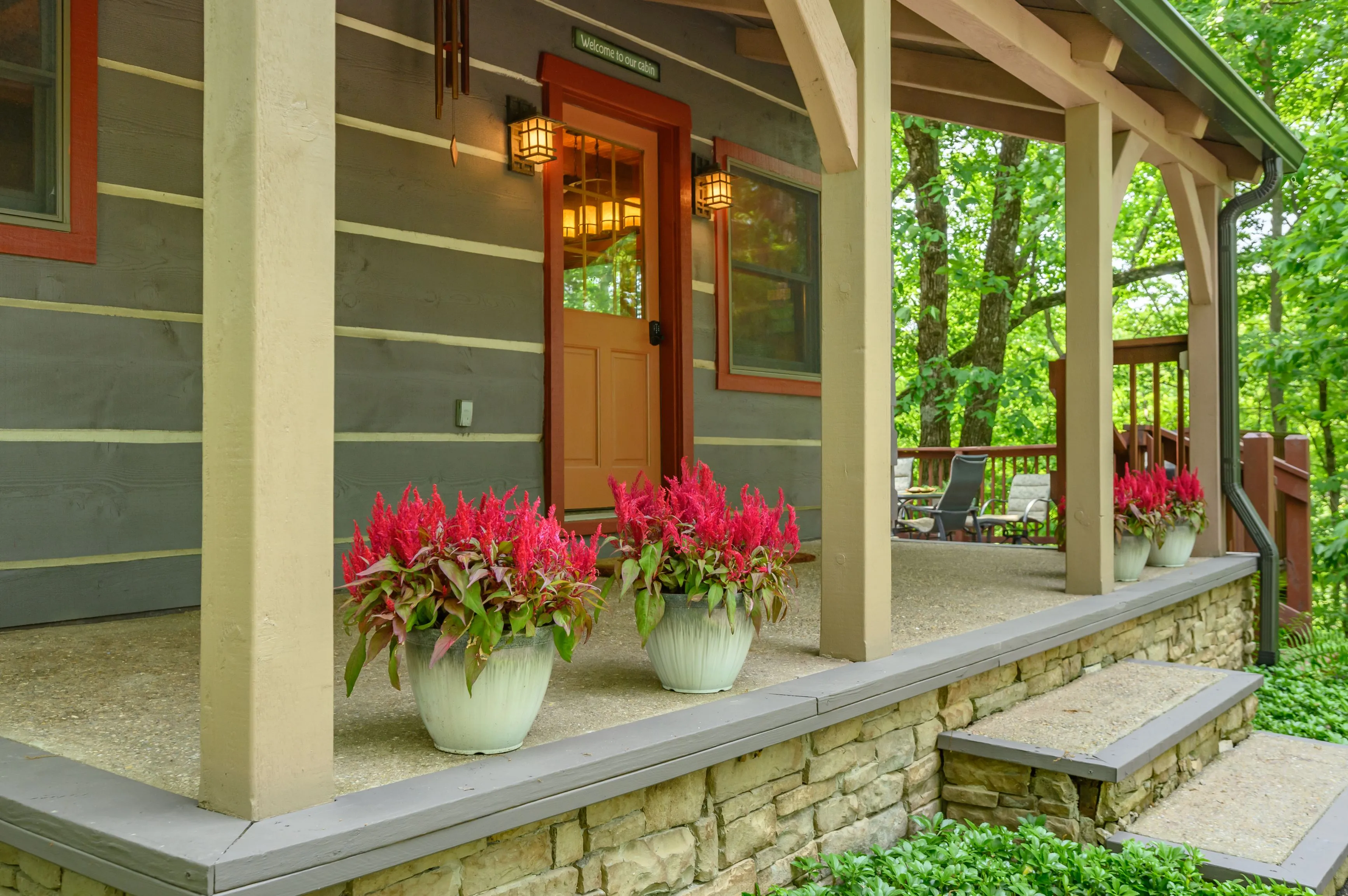 Exterior view of a cozy cottage with a porch, decorative red flowers in pots, and forest in the background.