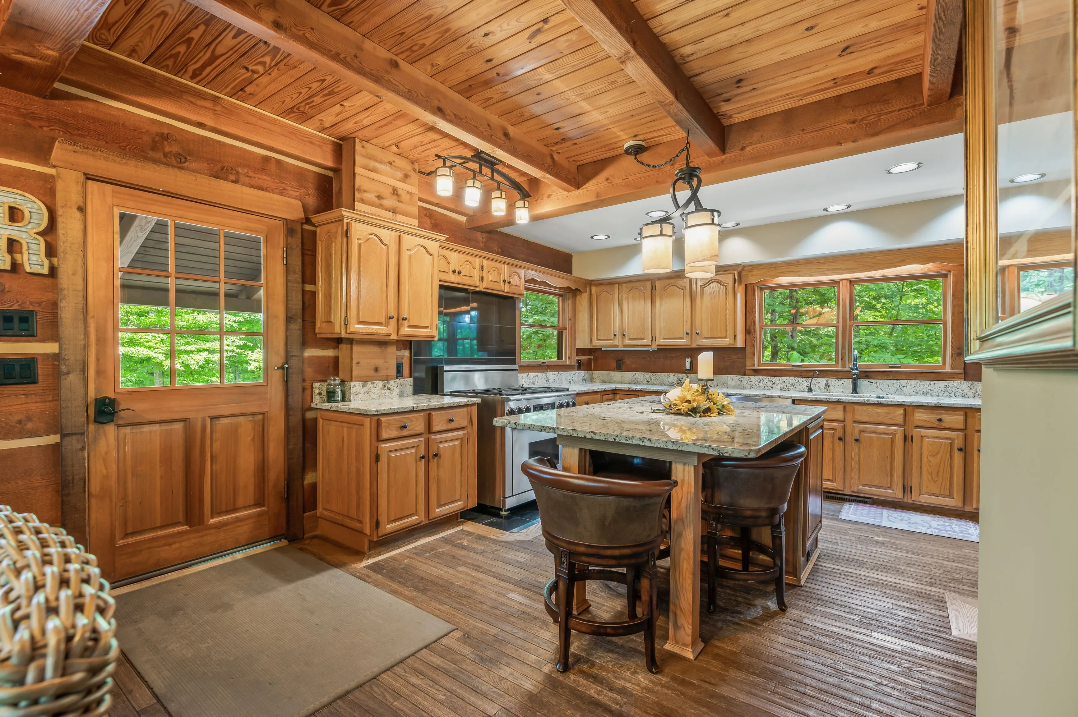 Spacious kitchen interior with wooden cabinets, granite countertops, and a central island with bar stools, under exposed wooden beams and modern lighting fixtures.