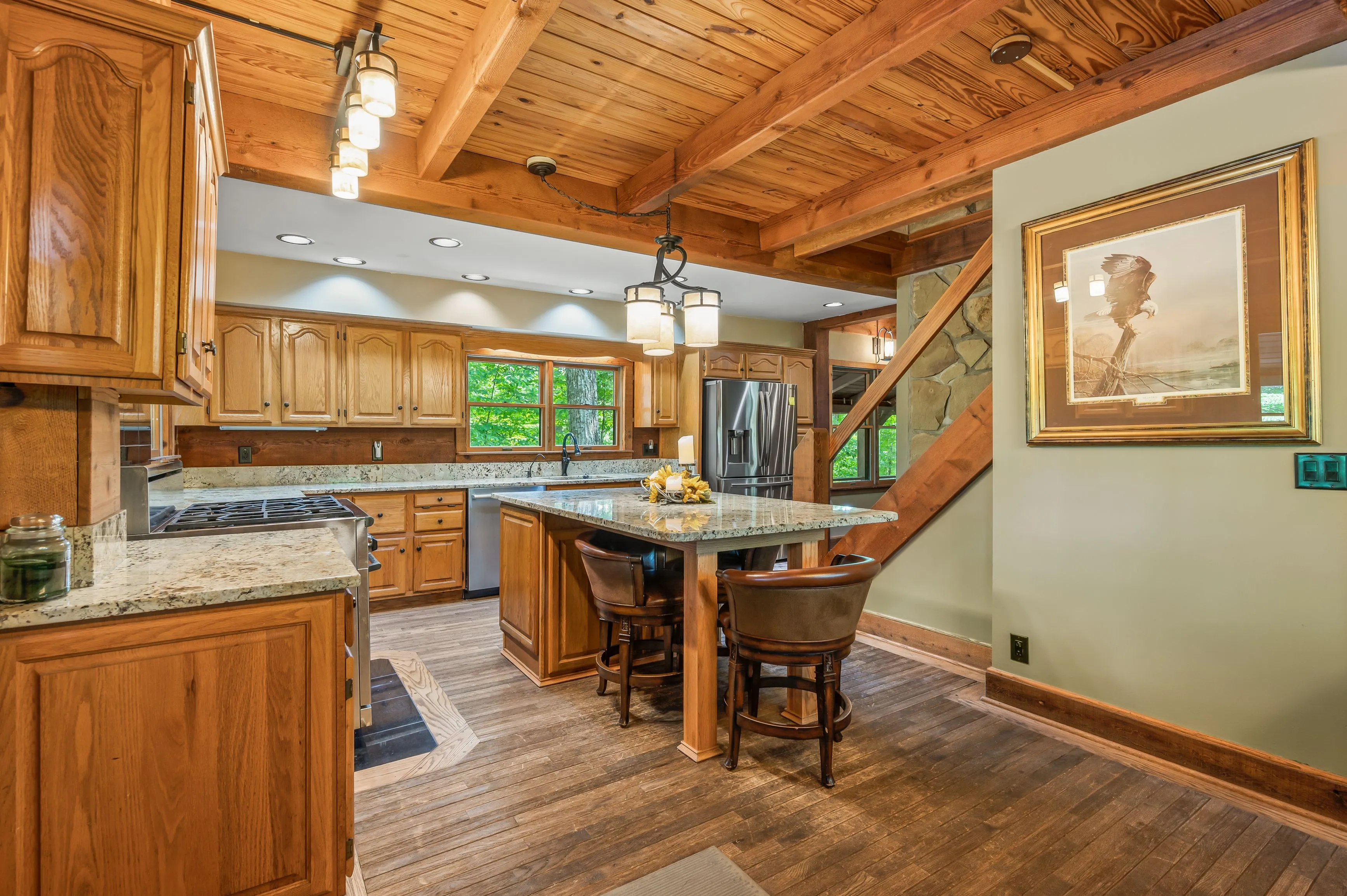 Spacious kitchen interior with wooden cabinets, granite countertops, and a central island with bar stools, featuring exposed wooden beams and a framed picture on the wall.