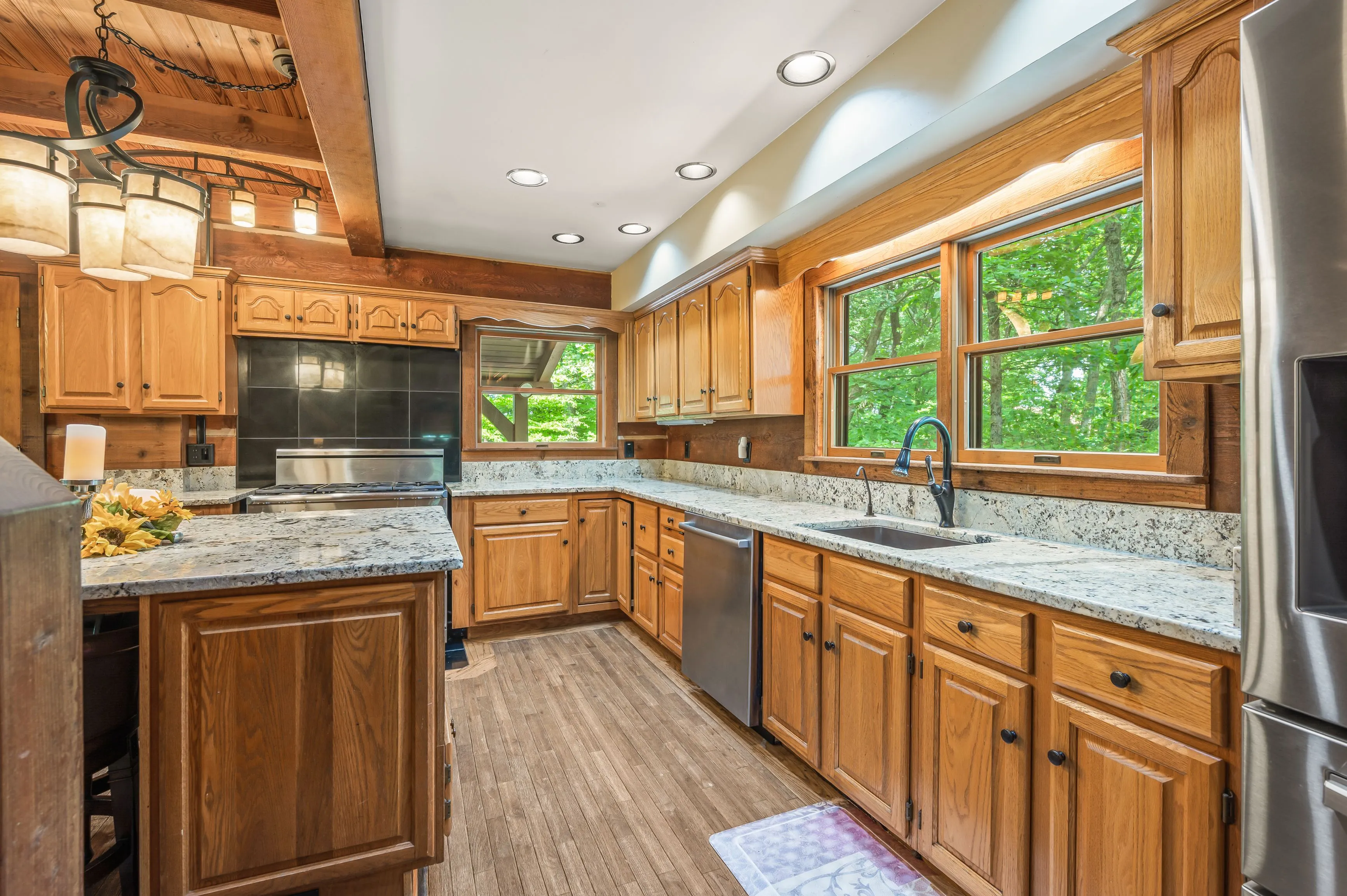 A bright, modern kitchen with wooden cabinets, granite countertops, and stainless steel appliances, surrounded by windows overlooking greenery.