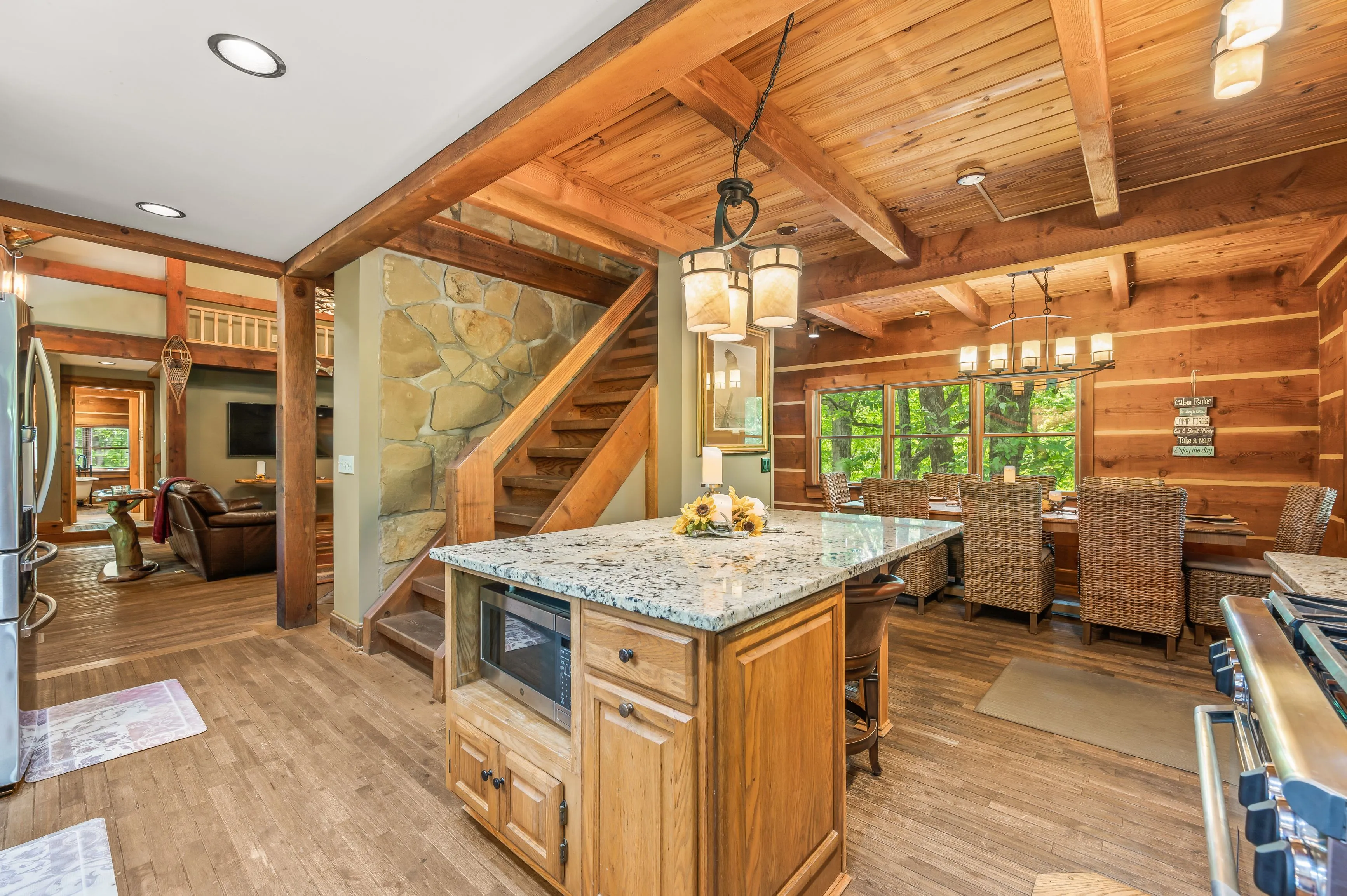Spacious rustic kitchen with wooden cabinetry, granite countertops, and stainless steel appliances, featuring exposed wooden beams, a stone staircase, and a dining area with large windows overlooking greenery.